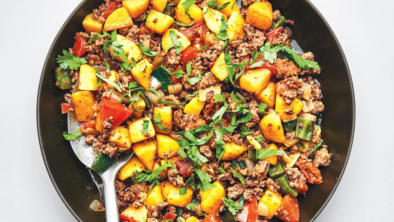 A cast iron skillet filled with Mexican picadillo, showing browned ground beef, tender potato cubes, tomatoes, and peas, garnished with cilantro and lime wedges on a warm kitchen counter