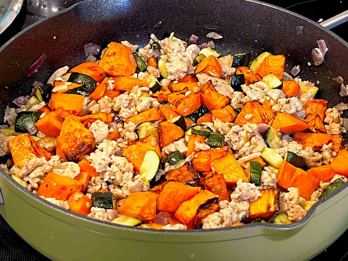 A cast iron skillet filled with browned chicken thighs, caramelized sweet potato chunks, red onion, and wilted greens, finished with a drizzle of creamy lime yogurt sauce on a bright kitchen counter