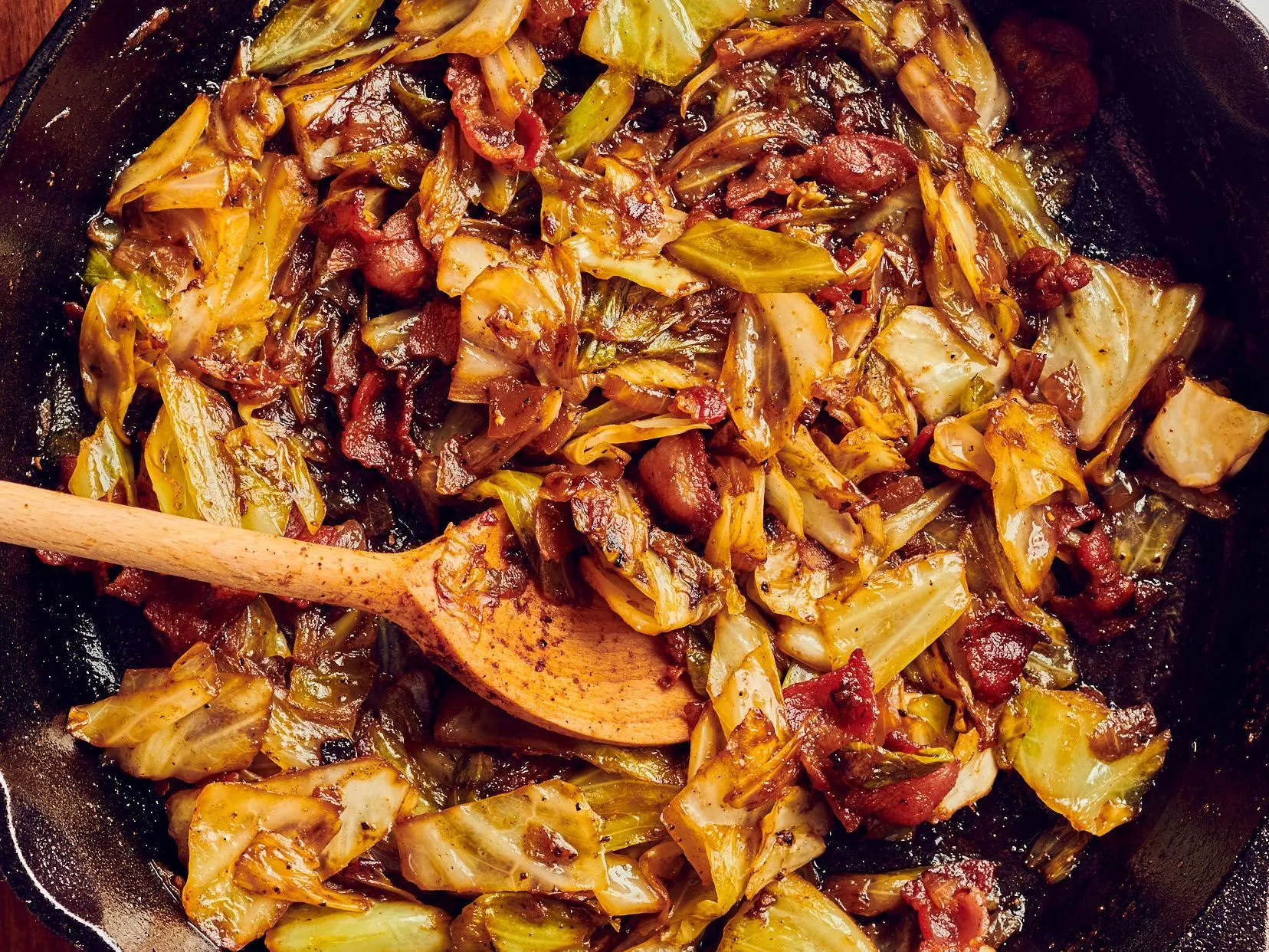 A cast iron skillet filled with caramelized green cabbage and bacon, with crisp browned edges and glossy butter, shot on a warm kitchen counter