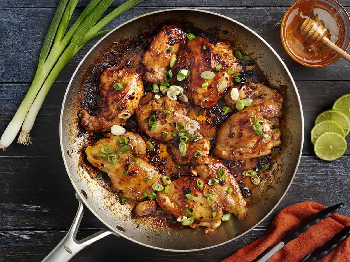 A cast iron skillet filled with chipotle honey glazed chicken thighs with caramelized edges, scattered cilantro, and lime wedges on a warm kitchen counter