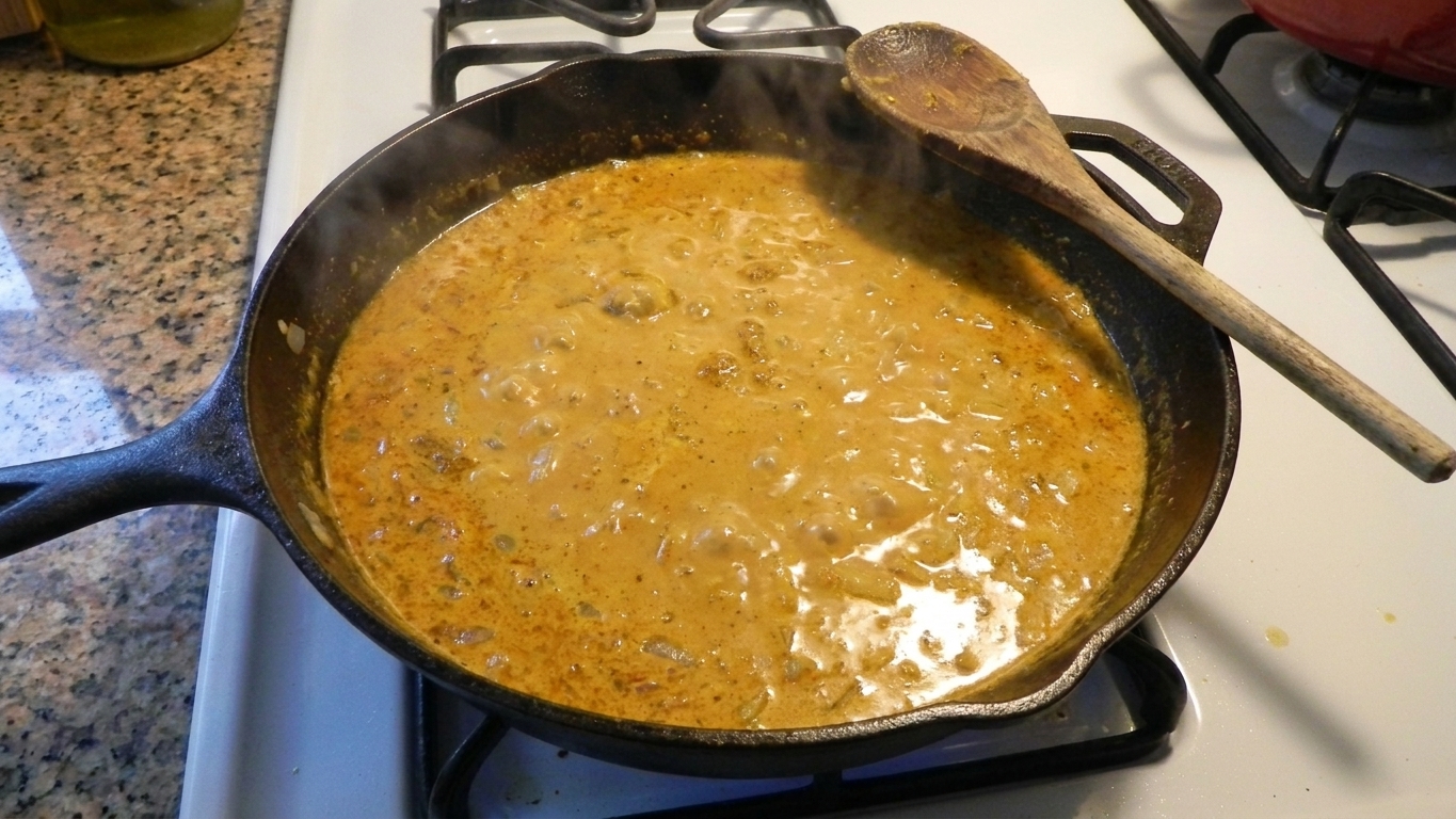 A cast iron skillet filled with creamy golden curry sauce, lightly bubbling on a stovetop with a wooden spoon resting on the edge