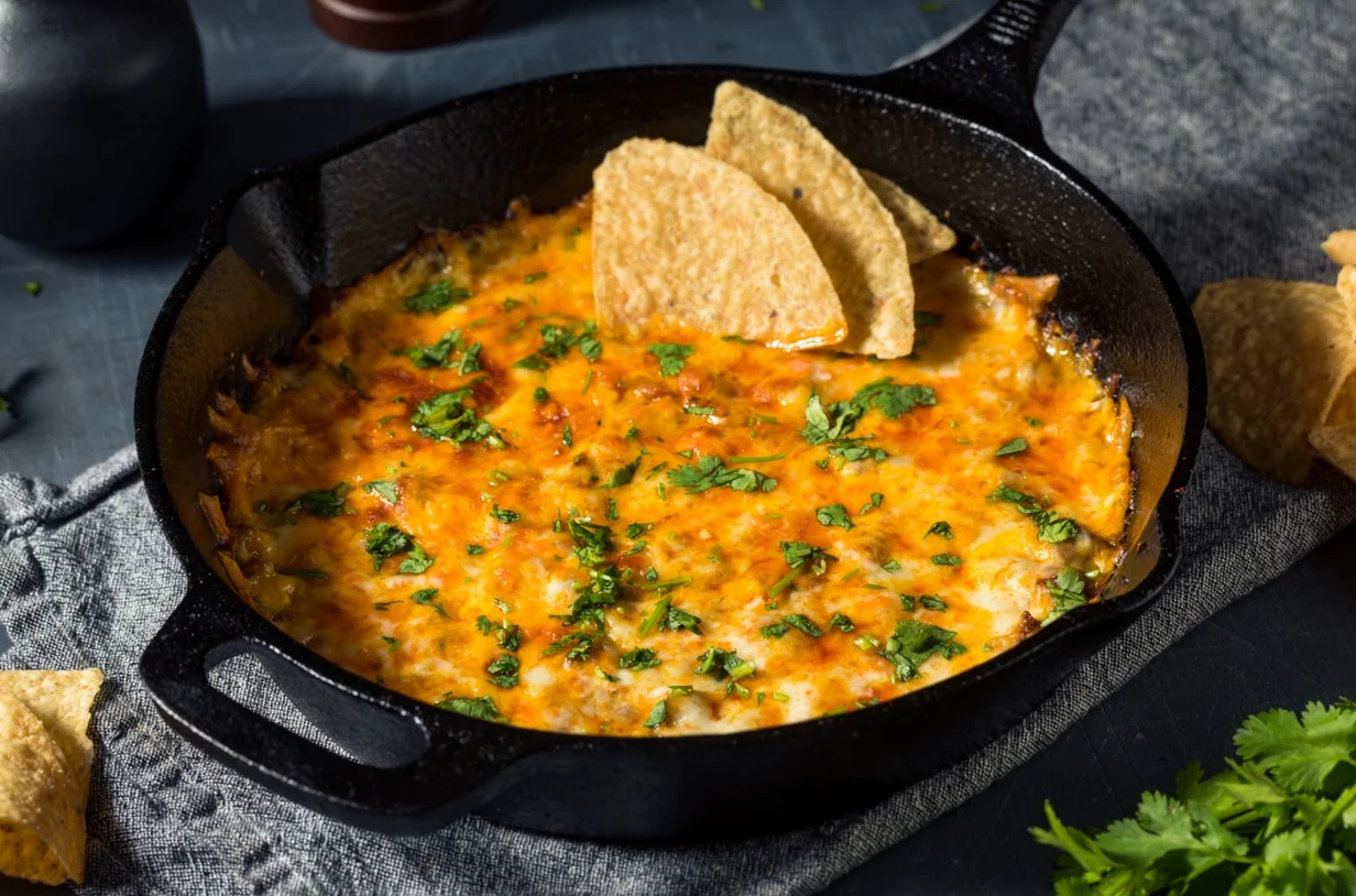 A cast iron skillet filled with creamy smoked queso topped with browned chorizo and diced cilantro, sitting on a wooden patio table with tortilla chips nearby