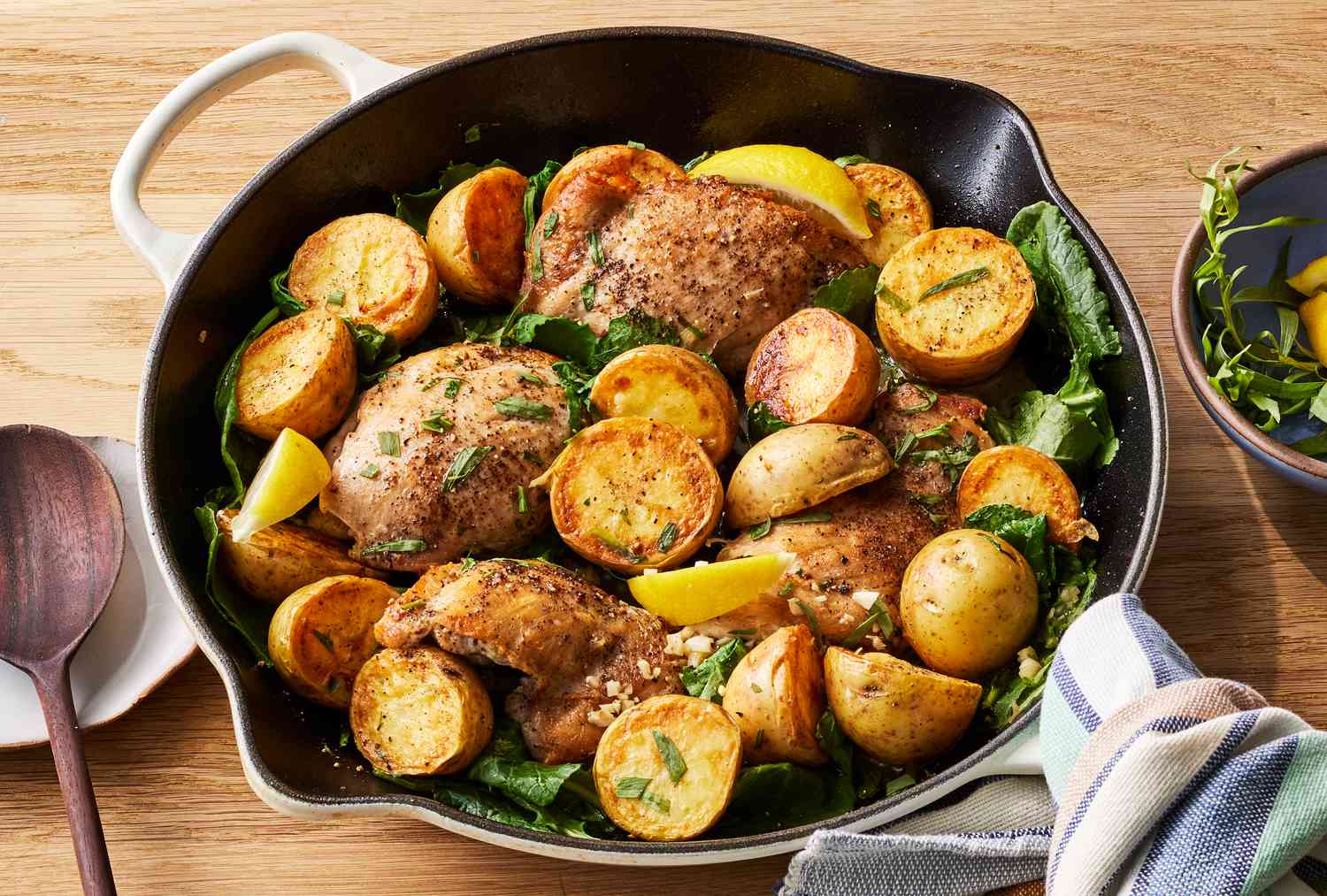 A cast iron skillet filled with crispy chicken thighs and golden roasted potato chunks, finished with lemon slices and chopped parsley on a warm kitchen counter