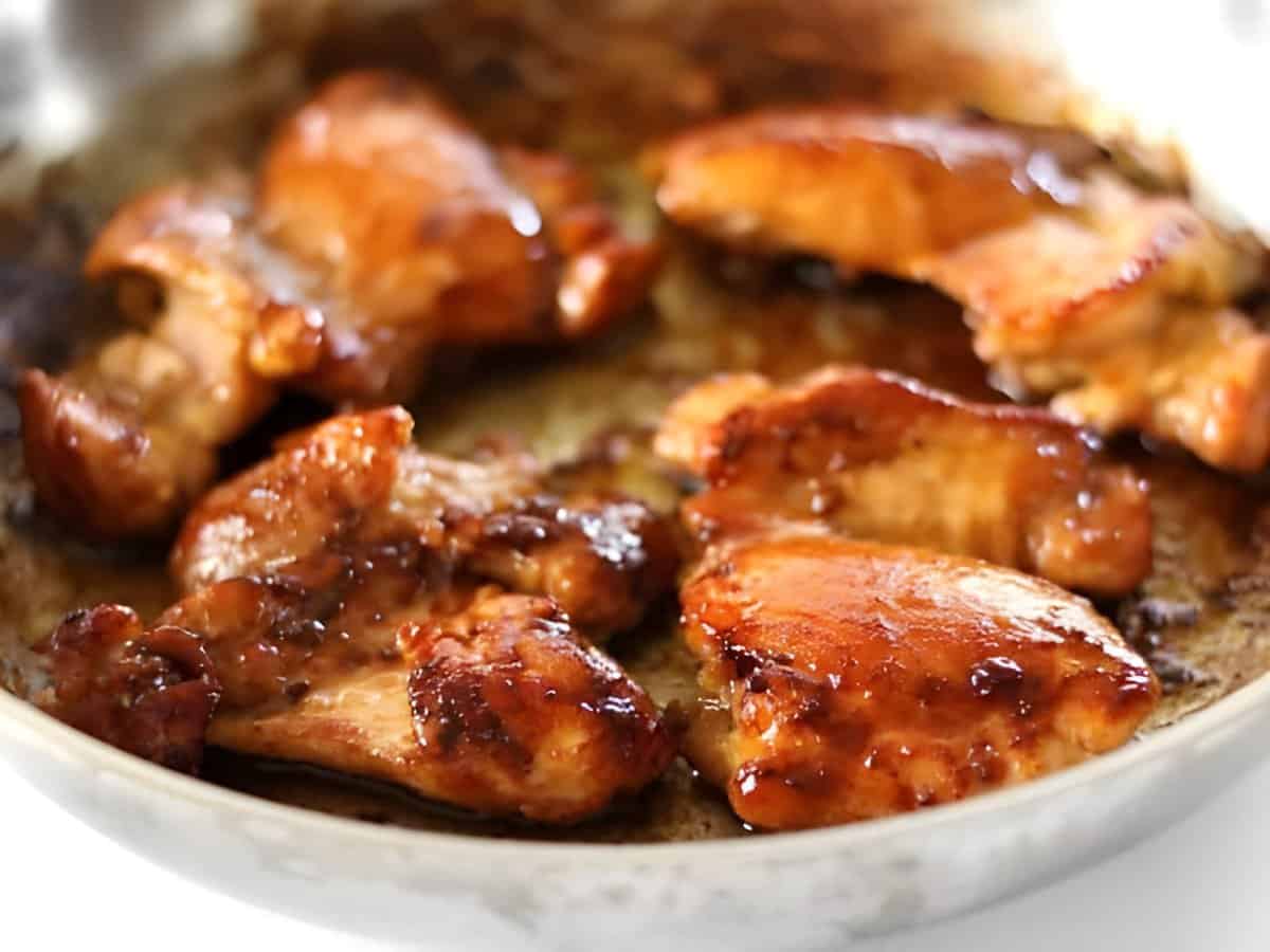 A cast iron skillet filled with glossy sticky ginger chicken pieces topped with sliced green onions and sesame seeds, with a small bowl of rice nearby on a warm kitchen counter
