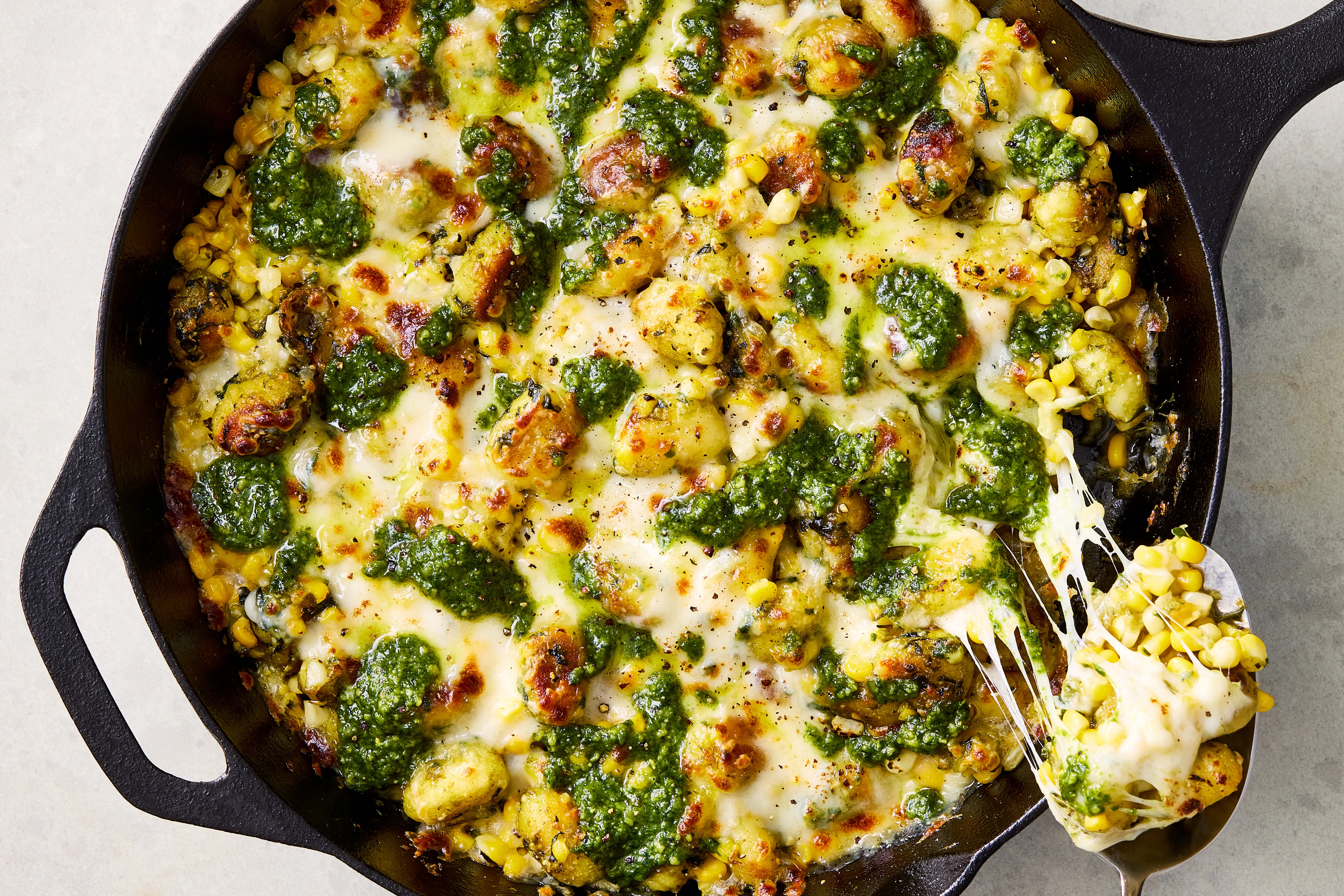 A cast iron skillet filled with golden crispy gnocchi, blistered cherry tomatoes, and melted mozzarella with pesto swirled throughout on a warm kitchen counter