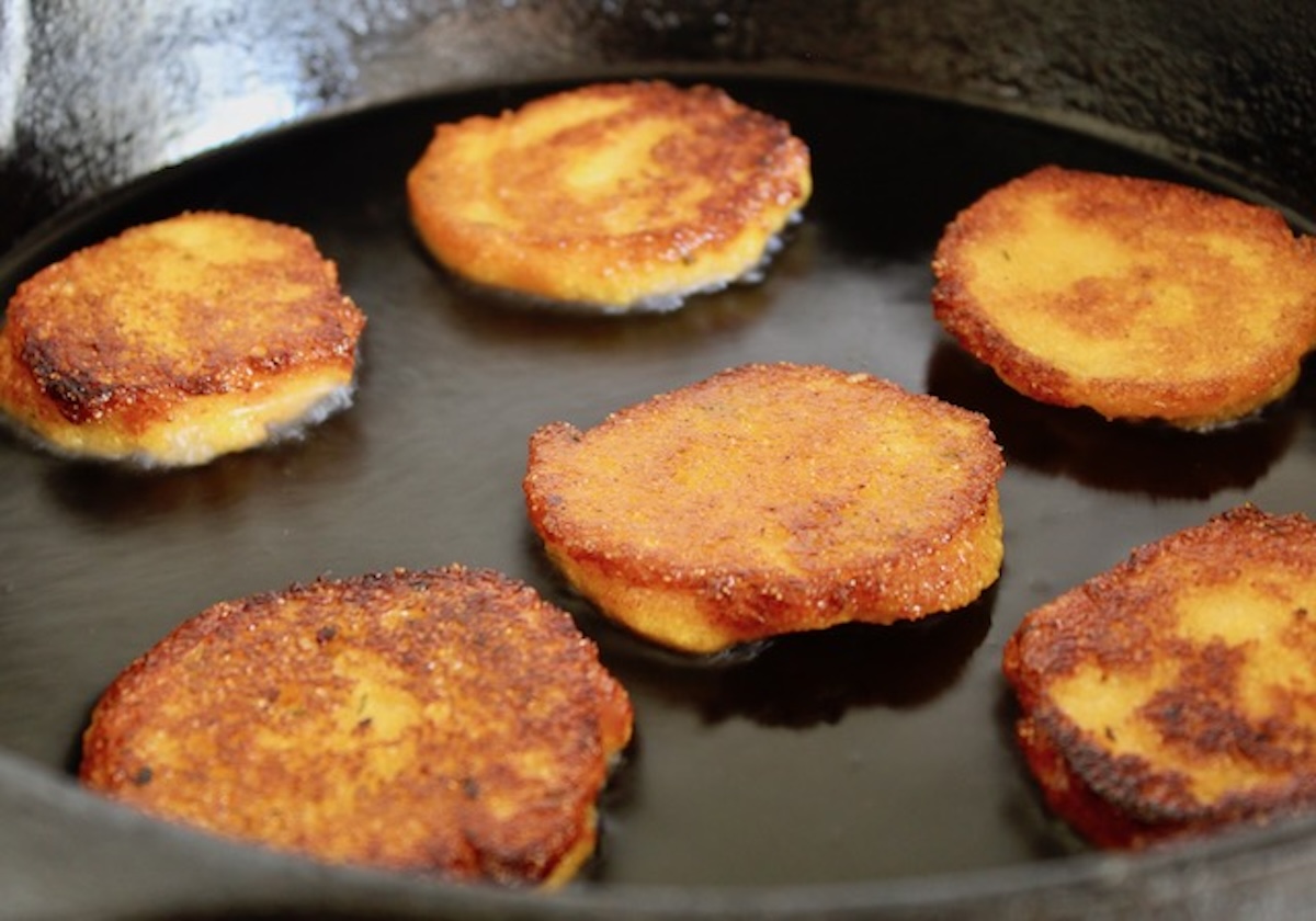 A cast iron skillet filled with golden hot water cornbread cakes frying in a shallow layer of oil, crisp edges visible on each cake