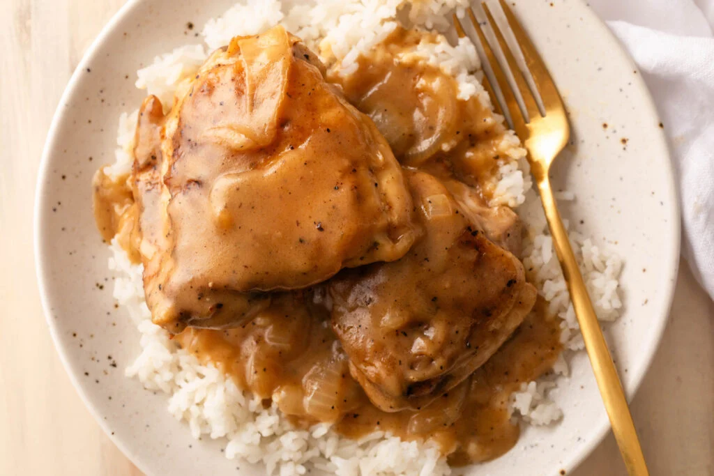 A cast iron skillet filled with golden seared chicken cutlets in peppery gravy, served with a bowl of buttery white rice on a wooden kitchen table in warm evening light