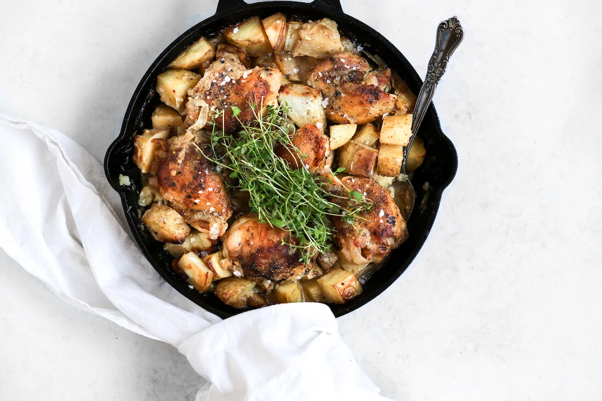 A cast iron skillet filled with golden seared chicken thighs coated in glossy honey mustard sauce, with chopped parsley and lemon wedges on a warm kitchen counter