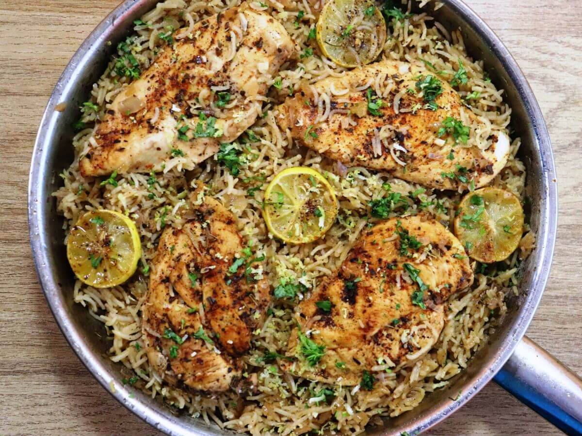 A cast iron skillet filled with golden seared chicken thighs nestled into fluffy rice with peas and lemon slices on a warm, neutral-toned kitchen counter, natural window light