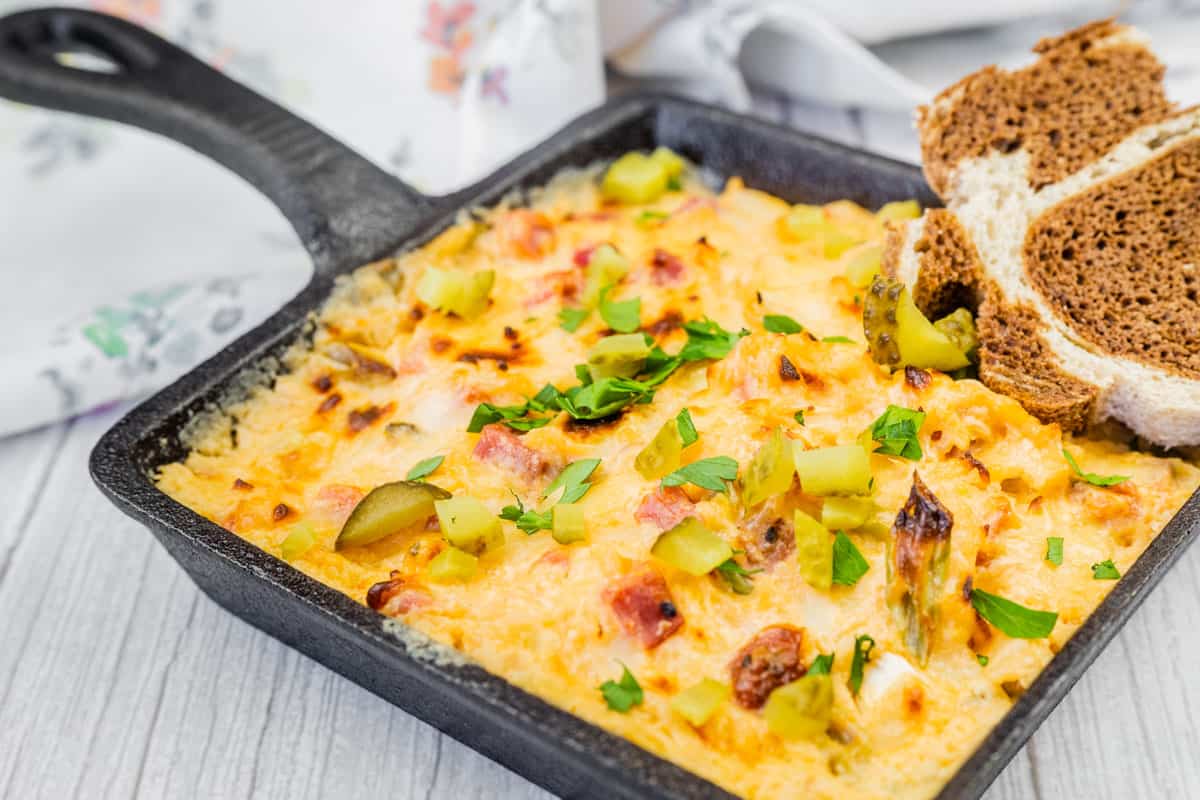 A cast iron skillet filled with hot Reuben dip, bubbling and browned on top with melted Swiss cheese and a drizzle of dressing, on a cozy kitchen counter with rye toasts nearby