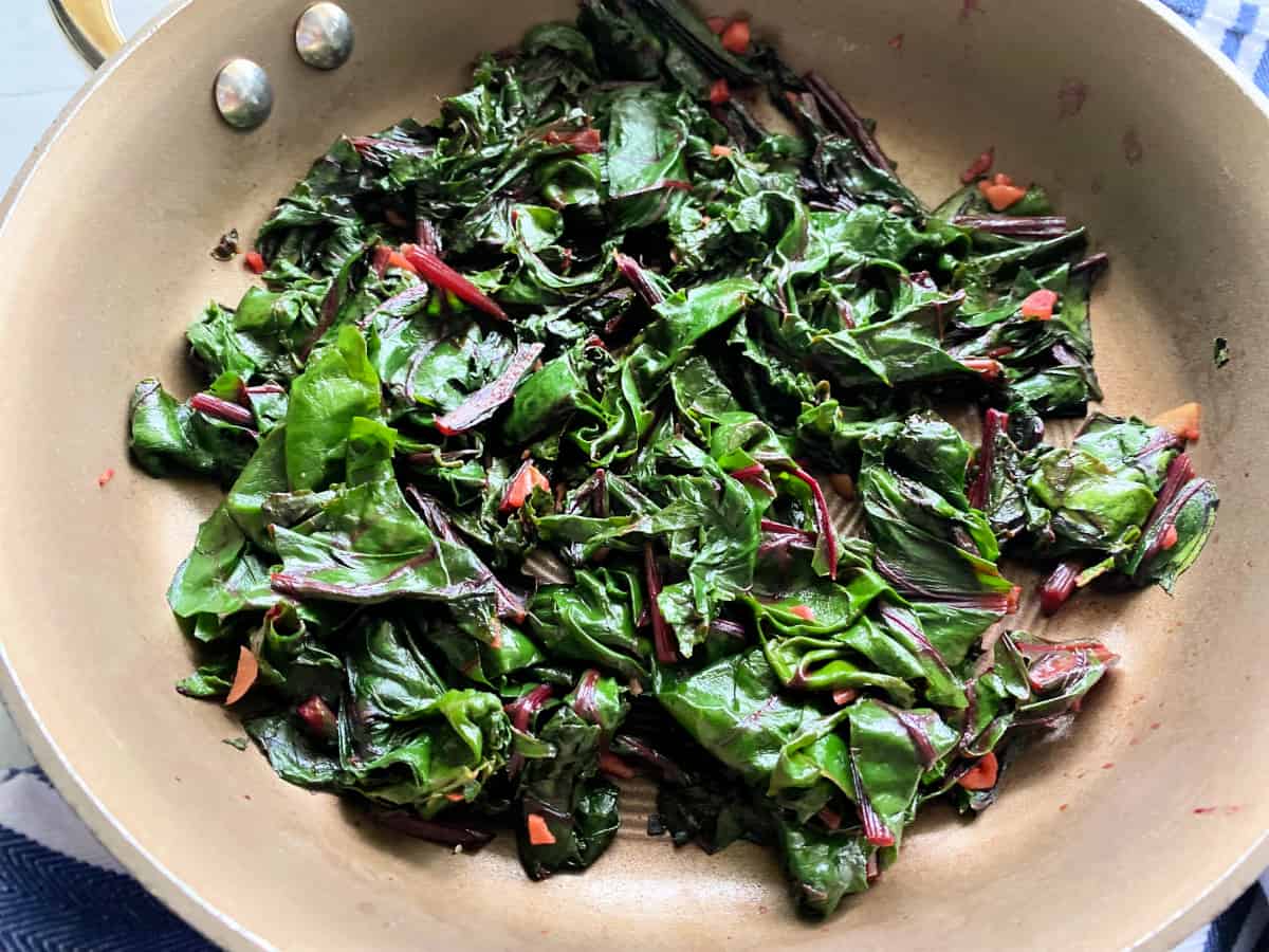 A cast iron skillet filled with sautéed beet greens and sliced stems, glossy with olive oil and garlic, with lemon wedges on a wooden cutting board beside it on a bright kitchen counter