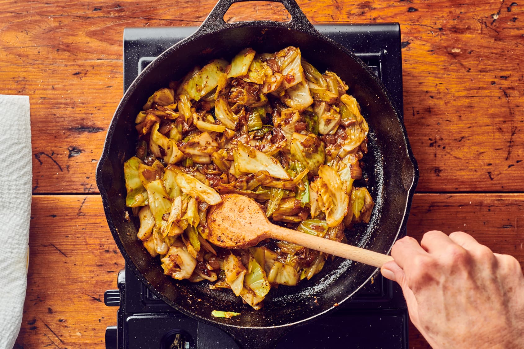 A cast iron skillet filled with sauteed green cabbage with golden brown edges and bits of garlic, set on a warm kitchen counter with a wooden spoon nearby