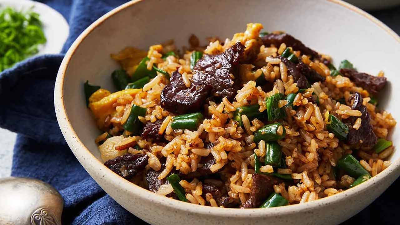 A cast iron skillet filled with steak fried rice, with browned rice, sliced leftover steak, scrambled egg, scallions, and sesame seeds
