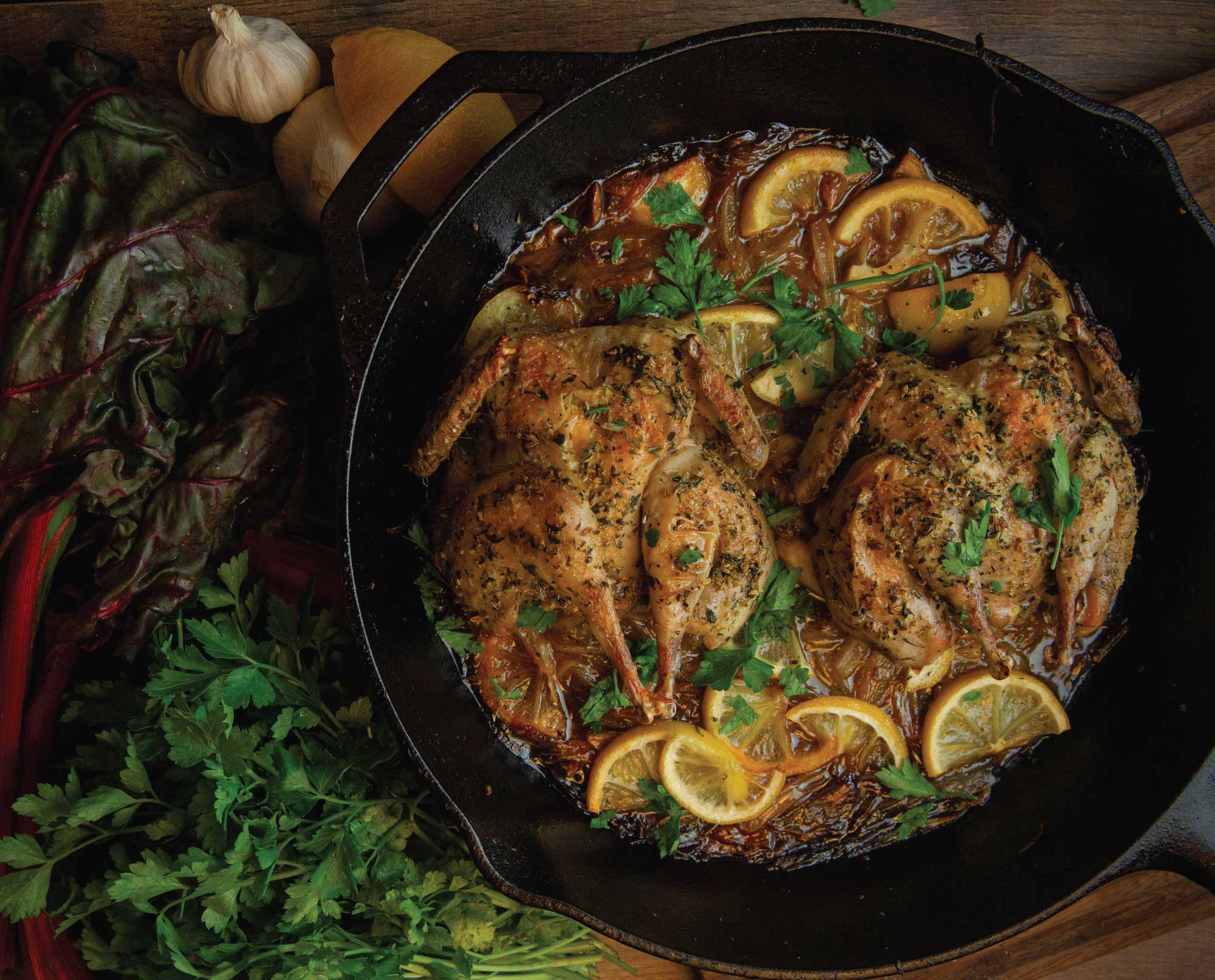 A cast iron skillet holding two browned, flattened quail with crispy skin, lemon halves, and a glossy pan sauce on a stovetop in a warm, home kitchen