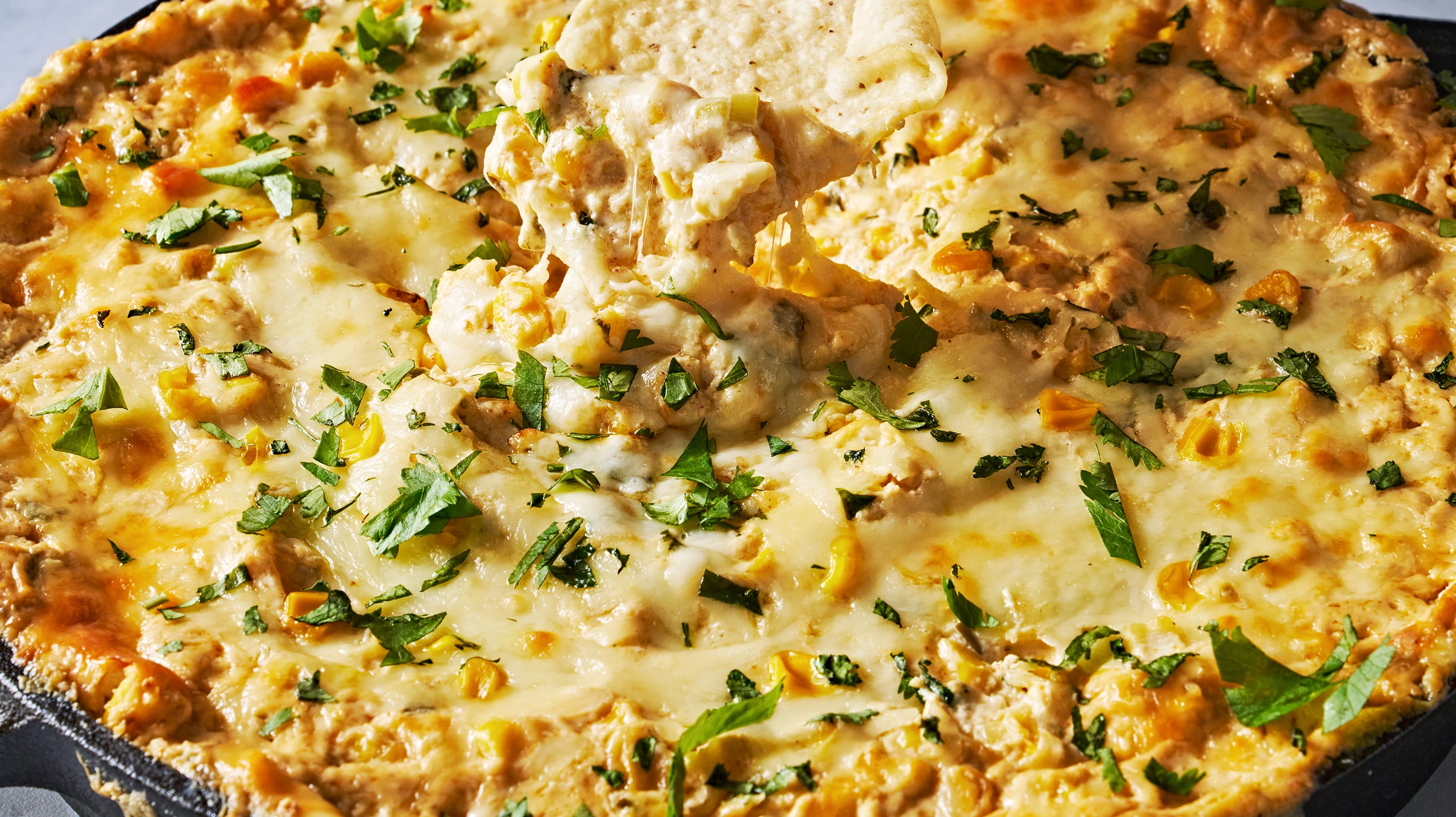 A cast iron skillet of baked Mexican corn dip, golden and bubbly on top with scattered cotija, cilantro, and a few jalapeño slices on a warm kitchen counter