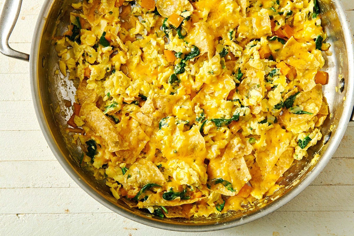 A cast iron skillet of migas with crispy tortilla chips, fluffy scrambled eggs, melted cheese, and chopped cilantro on a warm kitchen counter
