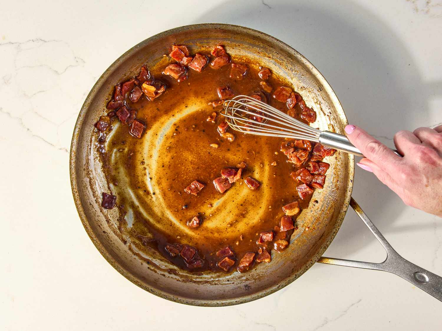 A cast iron skillet on a stovetop with glossy ham gravy being whisked, steam rising, with a wooden spoon and a small bowl of ham drippings nearby