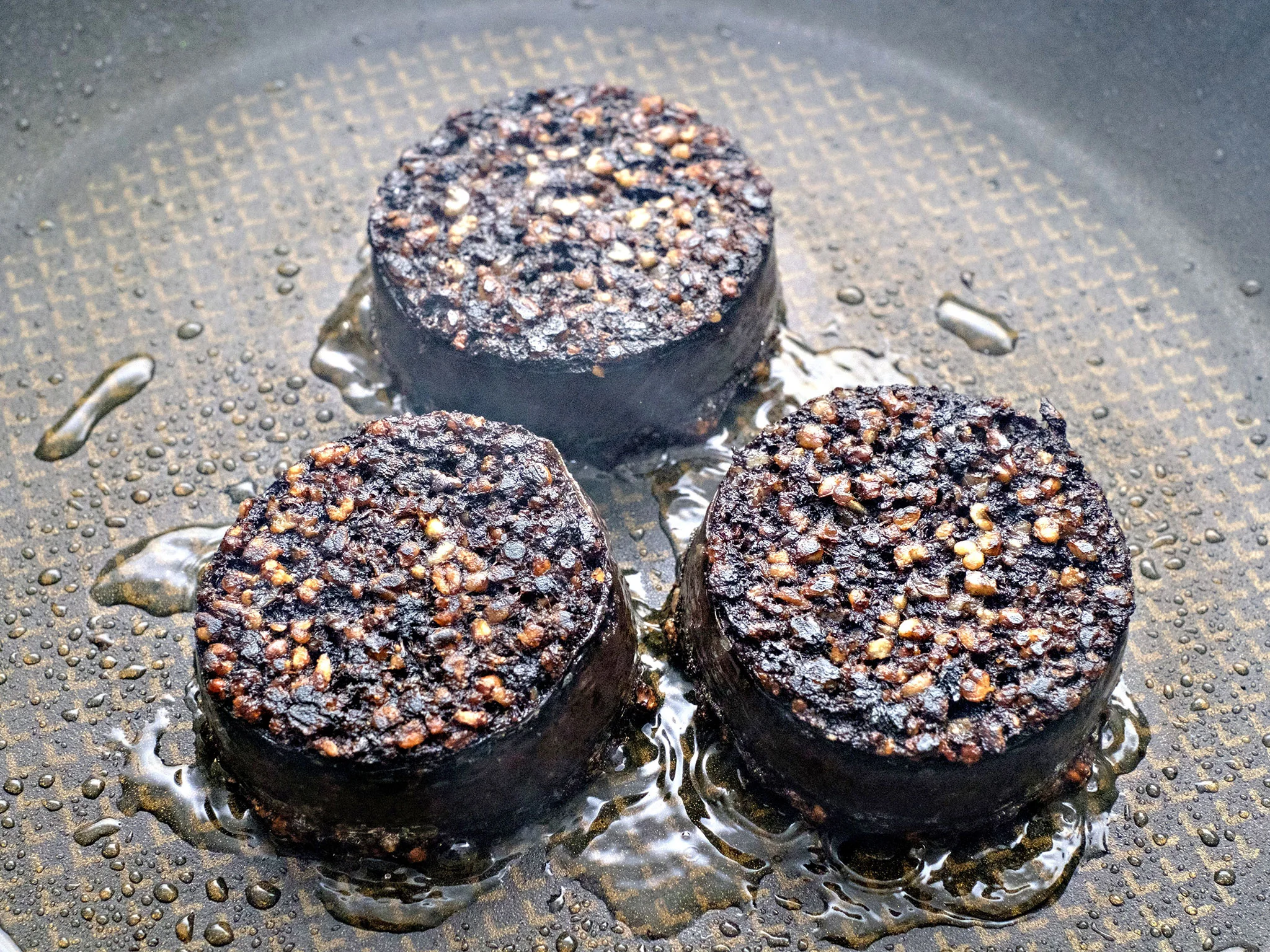 A cast iron skillet with thick slices of blood pudding sizzling and browning, with crisp edges forming, warm kitchen lighting