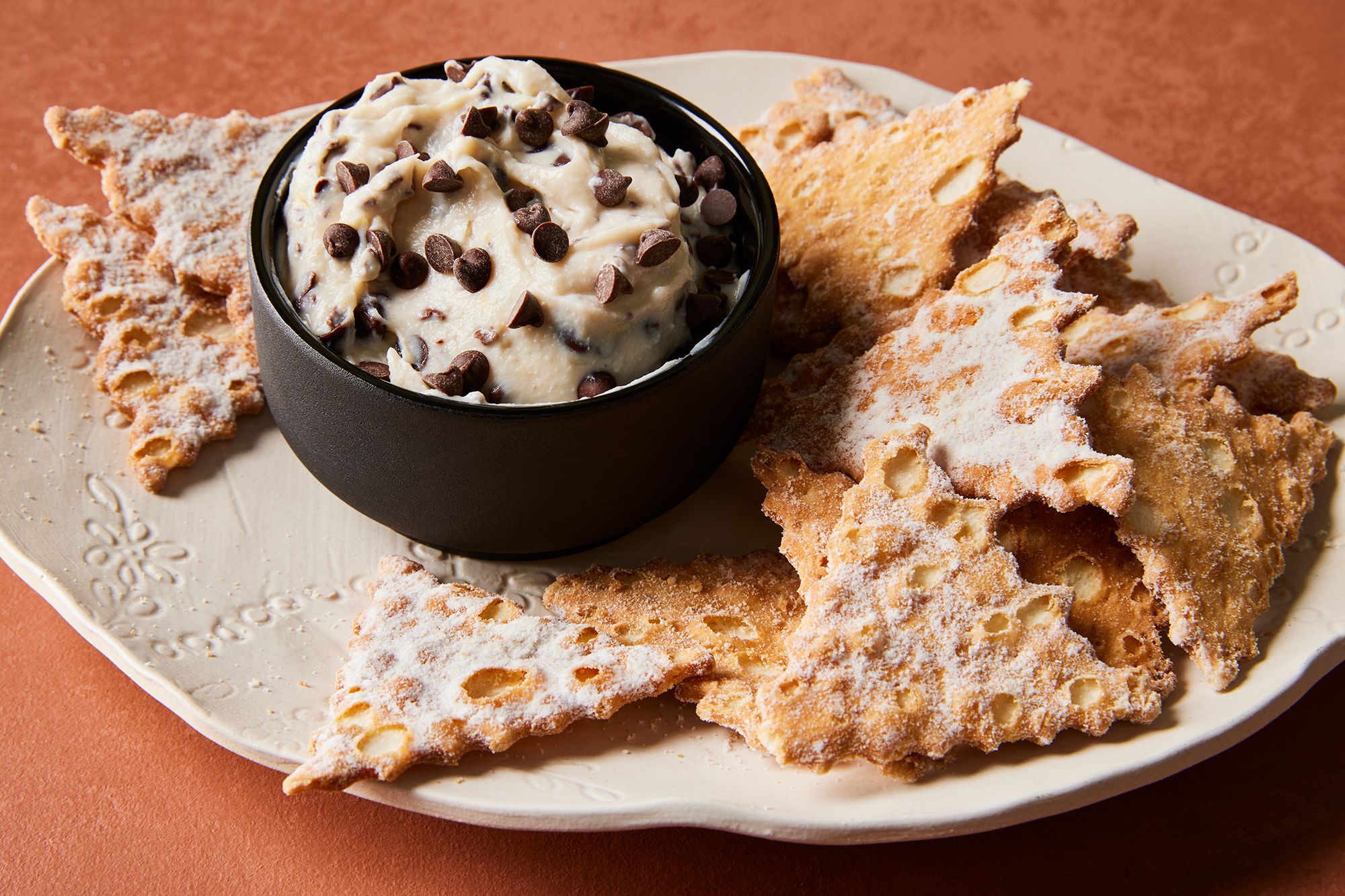 A ceramic bowl filled with creamy cannoli dip topped with mini chocolate chips and orange zest on a bright kitchen counter, surrounded by waffle cone pieces and berries