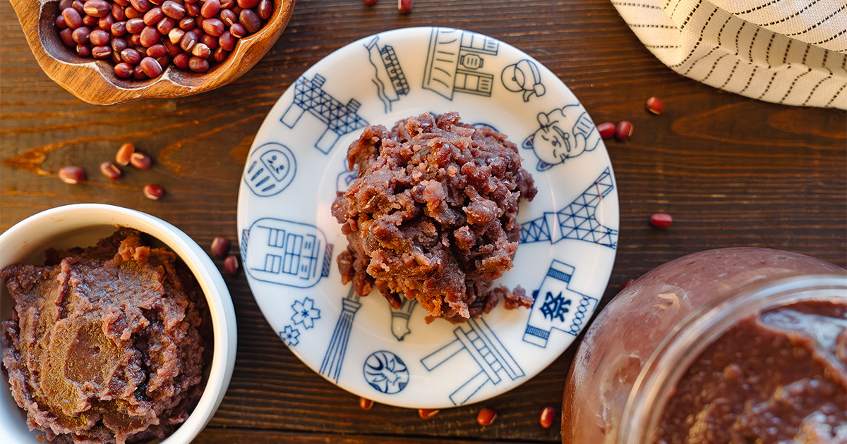 A ceramic bowl filled with glossy, smooth red bean paste on a wooden kitchen counter with a small spoon beside it