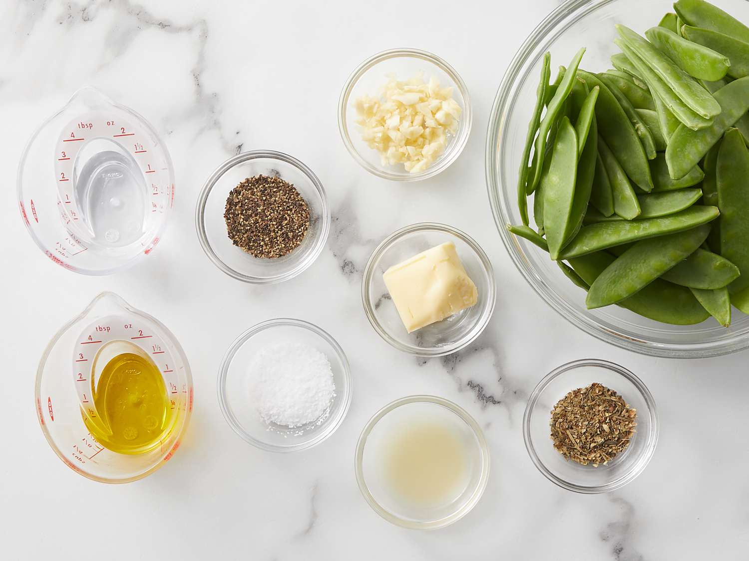 A ceramic bowl filled with trimmed snow peas on a kitchen counter beside minced garlic, soy sauce, butter, sesame oil, and a halved lemon