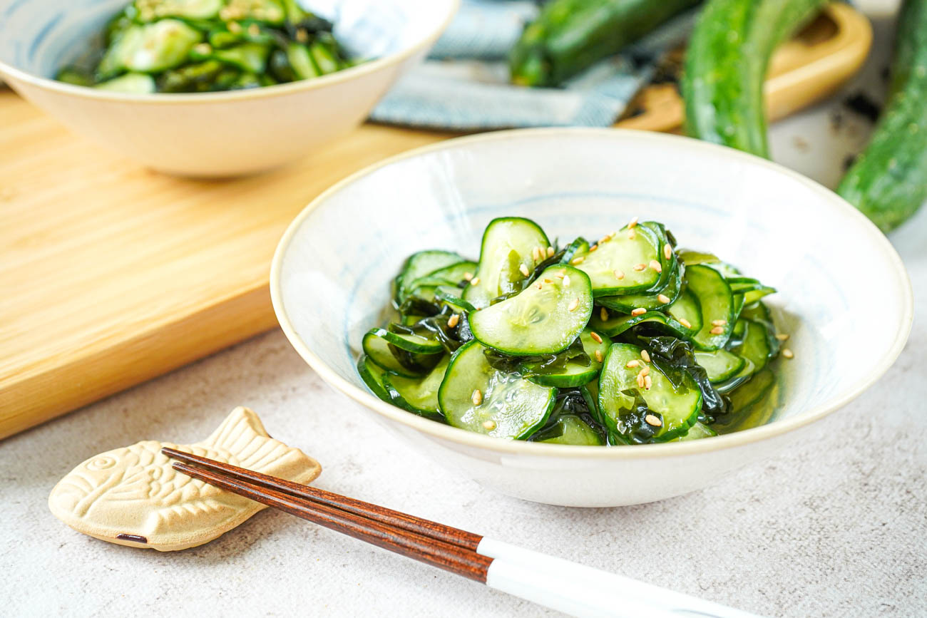 A ceramic bowl of Japanese cucumber salad with thin cucumber slices, sesame seeds, and a light vinegar dressing on a bright kitchen counter