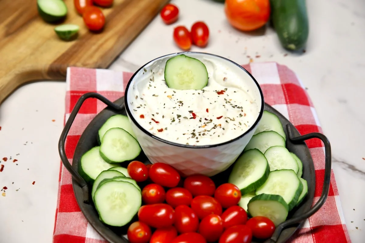 A ceramic bowl of creamy Greek yogurt veggie dip topped with chopped dill, black pepper, and a drizzle of olive oil, surrounded by carrots, cucumbers, and bell peppers on a light stone countertop