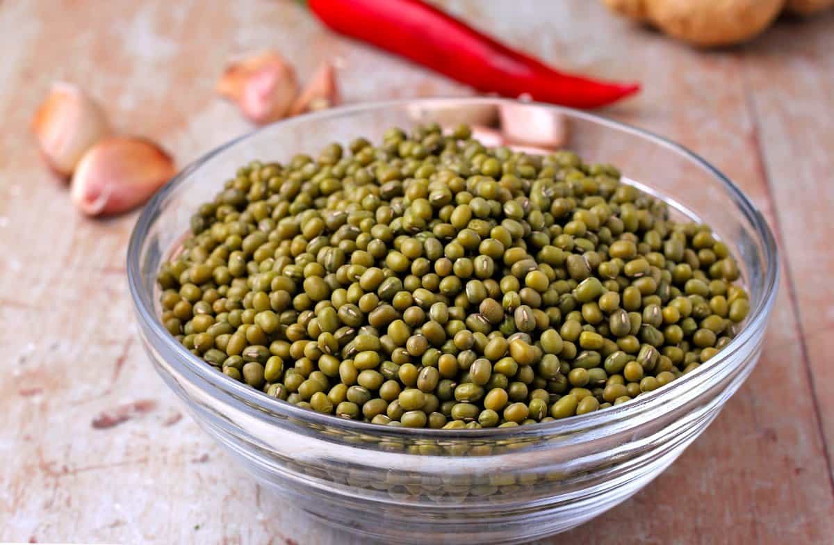 A ceramic bowl of dry green mung beans on a kitchen counter beside small ramekins of spices