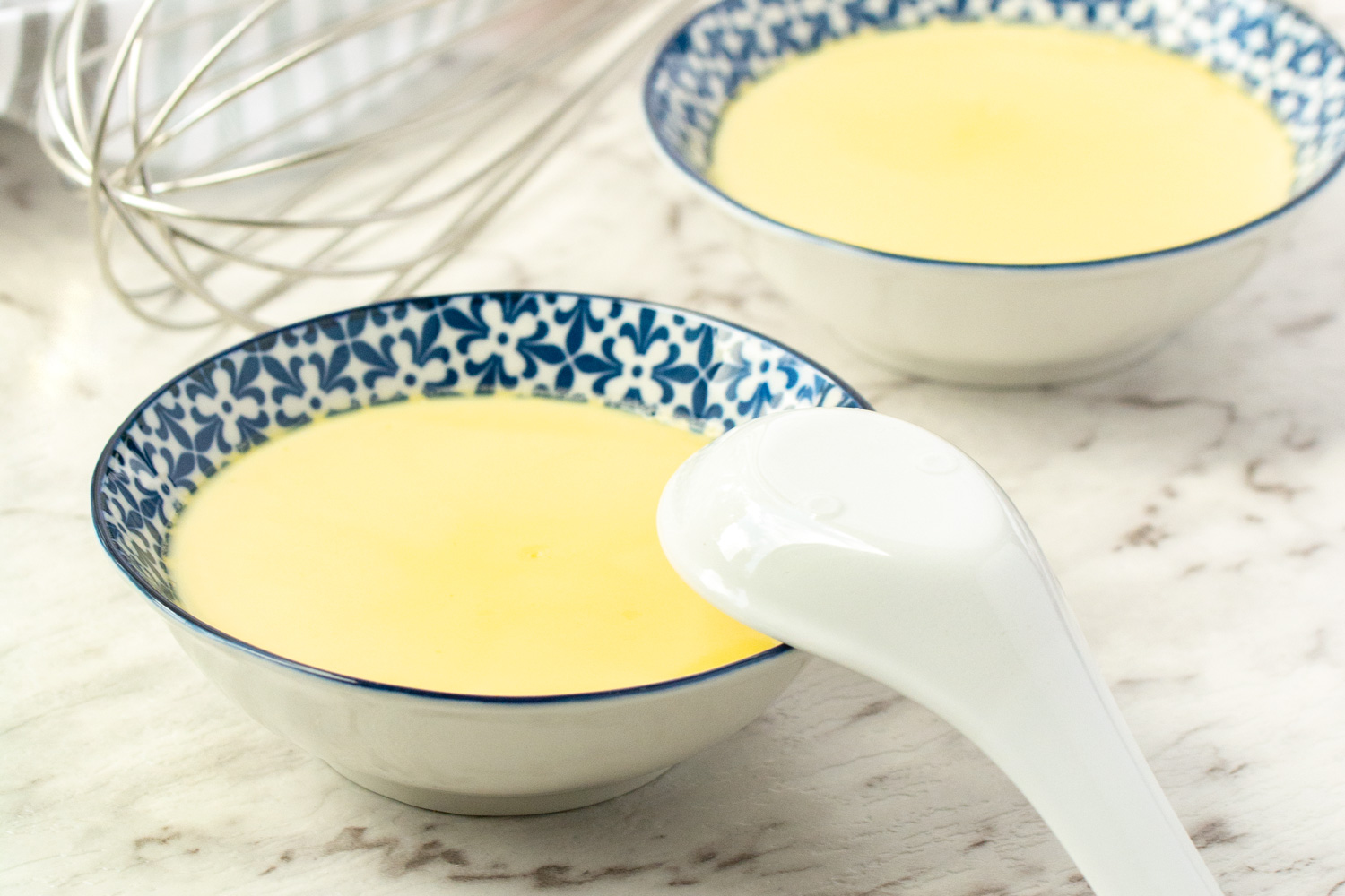 A ceramic bowl of silky Chinese steamed egg custard topped with sliced scallions and a drizzle of soy sauce on a light kitchen table