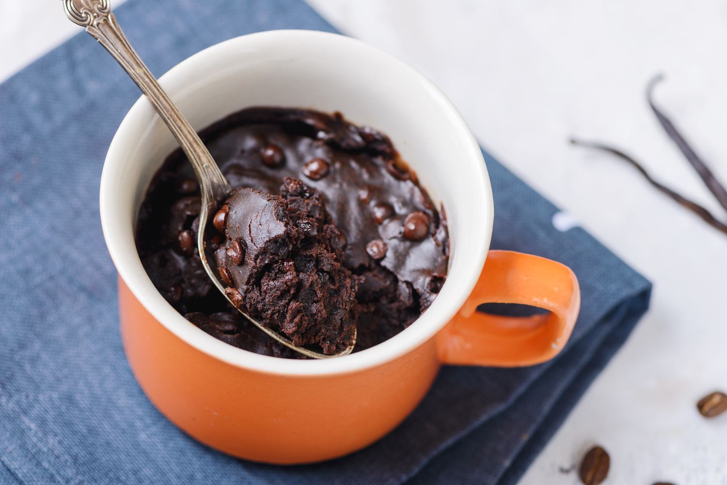 A ceramic mug filled with brownie batter sitting on a microwave turntable mid cook with the door open and a spoon resting on the counter nearby