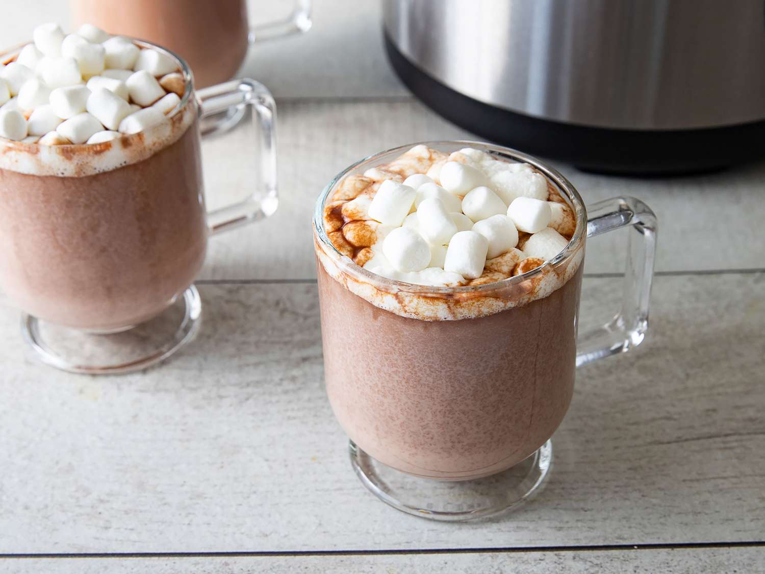 A ceramic mug of rich hot chocolate topped with whipped cream and chocolate shavings sitting beside a slow cooker on a warm kitchen counter with soft evening light