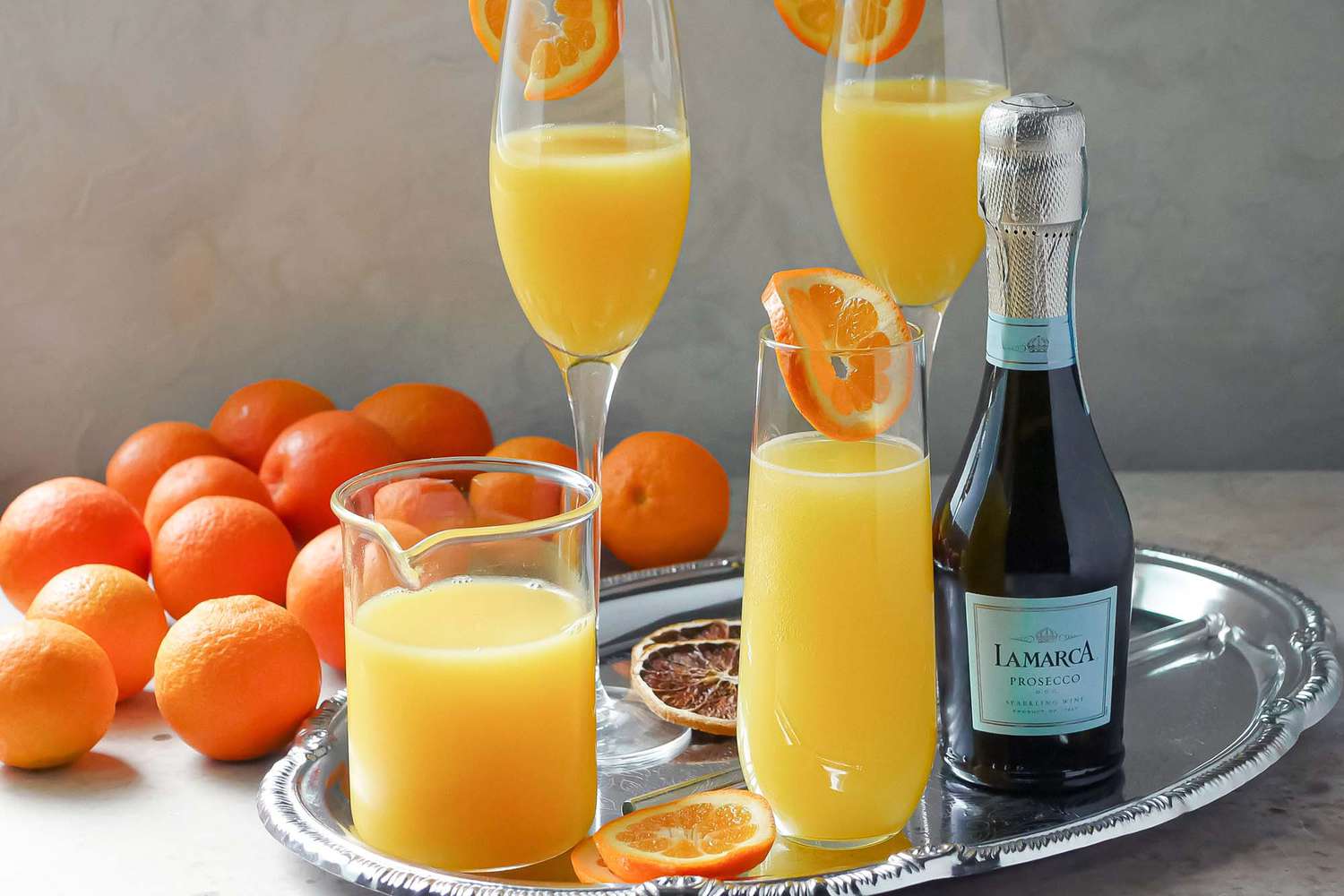 A chilled bottle of Brut Prosecco next to a pitcher of orange juice and sliced oranges on a bright kitchen counter