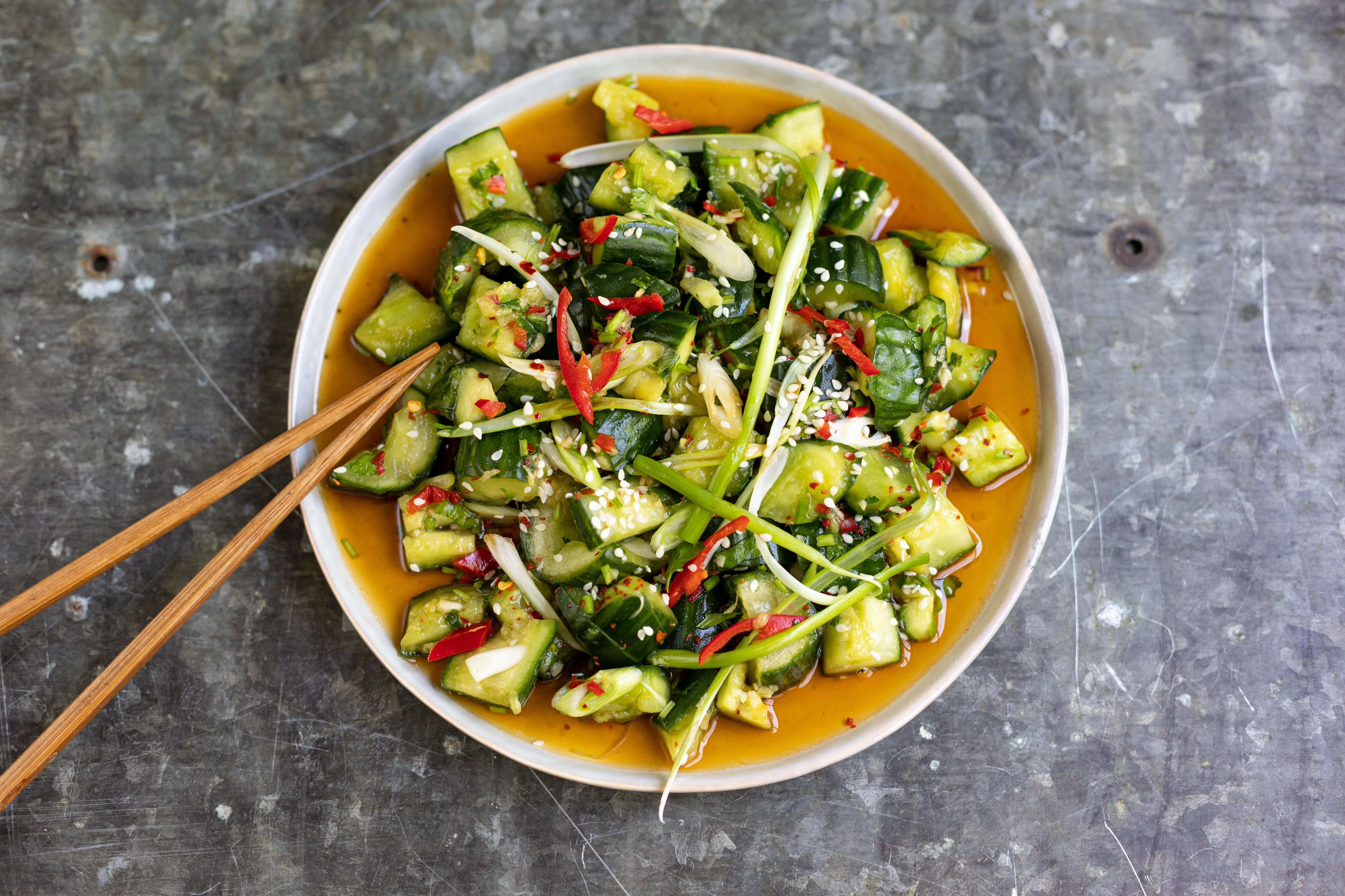 A chilled bowl of Din Tai Fung style cucumber salad with smashed cucumber chunks coated in a glossy garlicky soy dressing, sprinkled with sesame seeds on a light wooden table