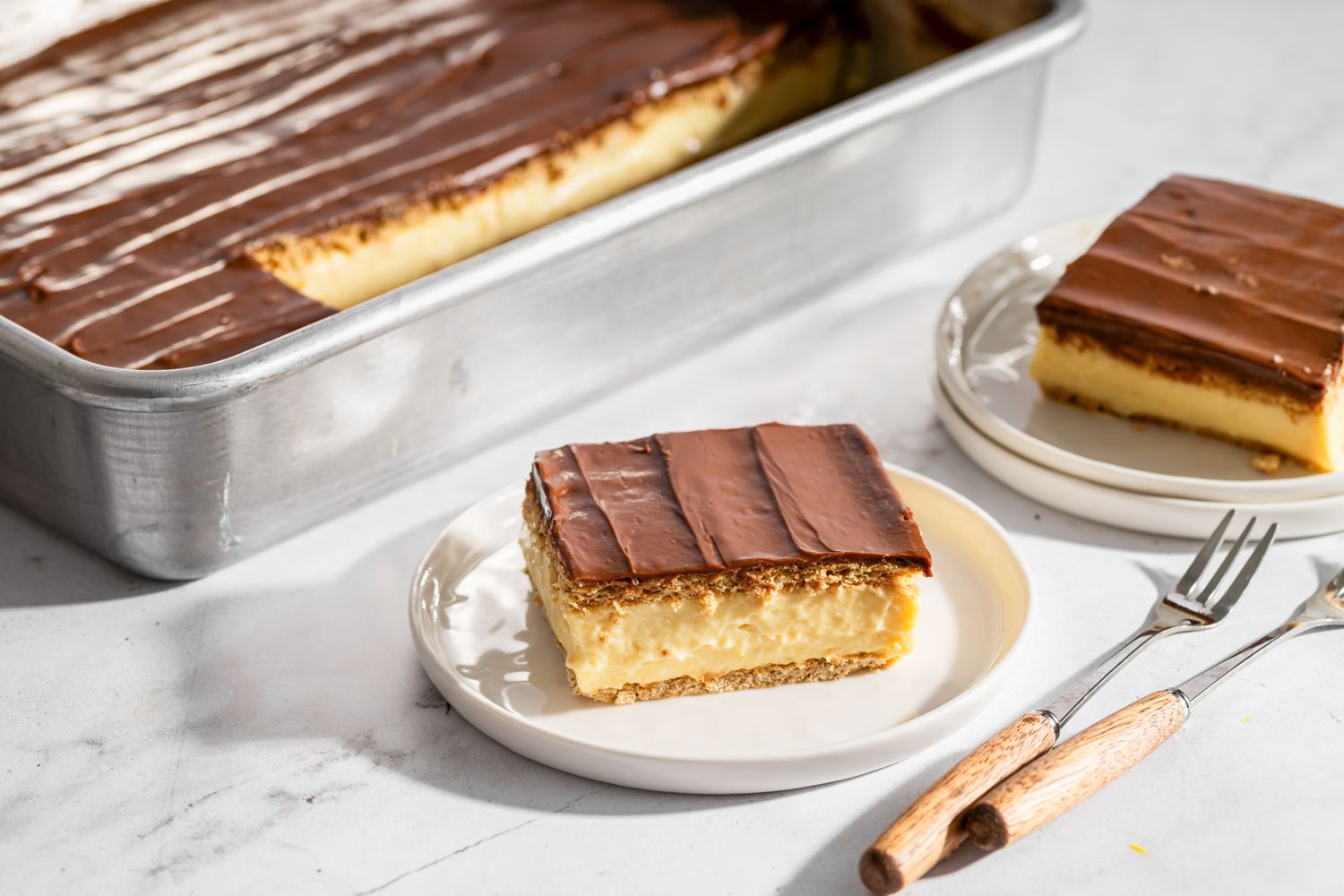 A chilled chocolate eclair cake in a glass 9x13 pan with one neat square slice removed, showing graham cracker layers and creamy vanilla filling under a glossy chocolate top, on a bright kitchen counter