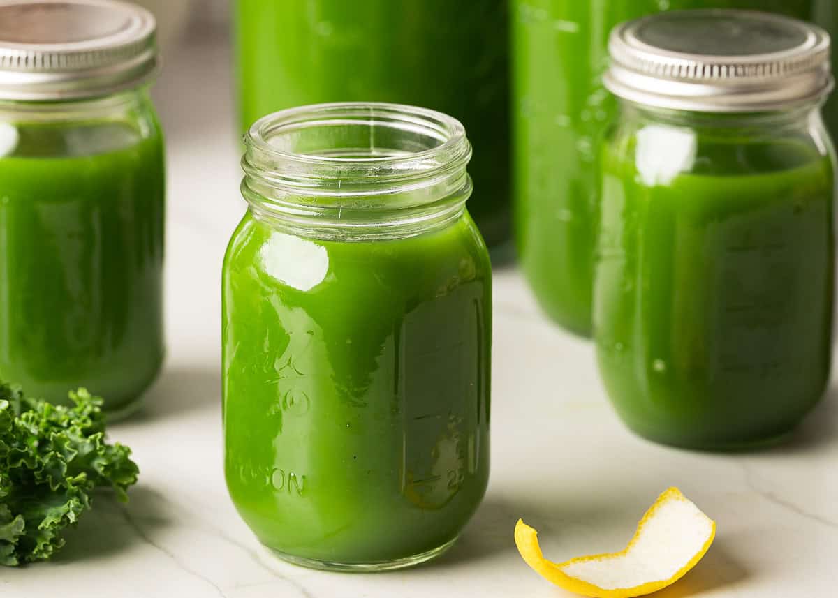 A chilled green juice in a clear mason jar on a bright kitchen counter with fresh cucumber, apple, lemon, and spinach nearby