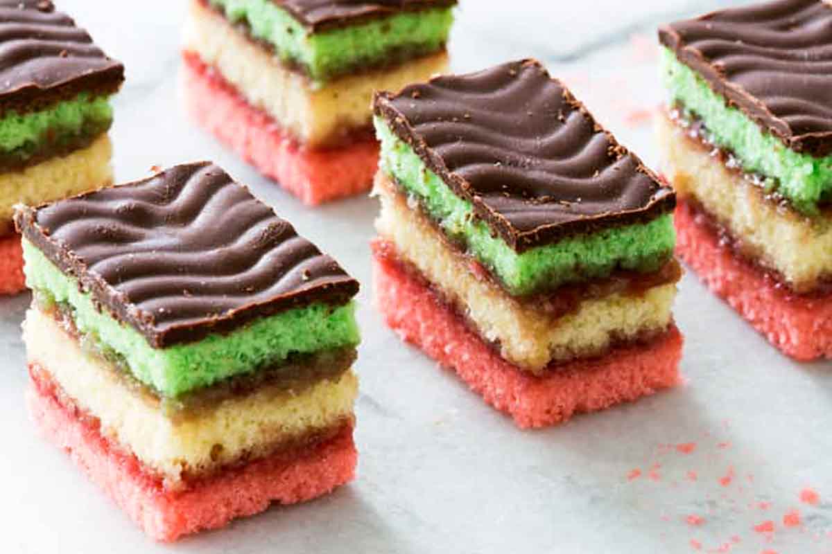 A chilled slab of Italian rainbow cookies being sliced into neat rectangles with a long knife on a cutting board, chocolate top glossy and set