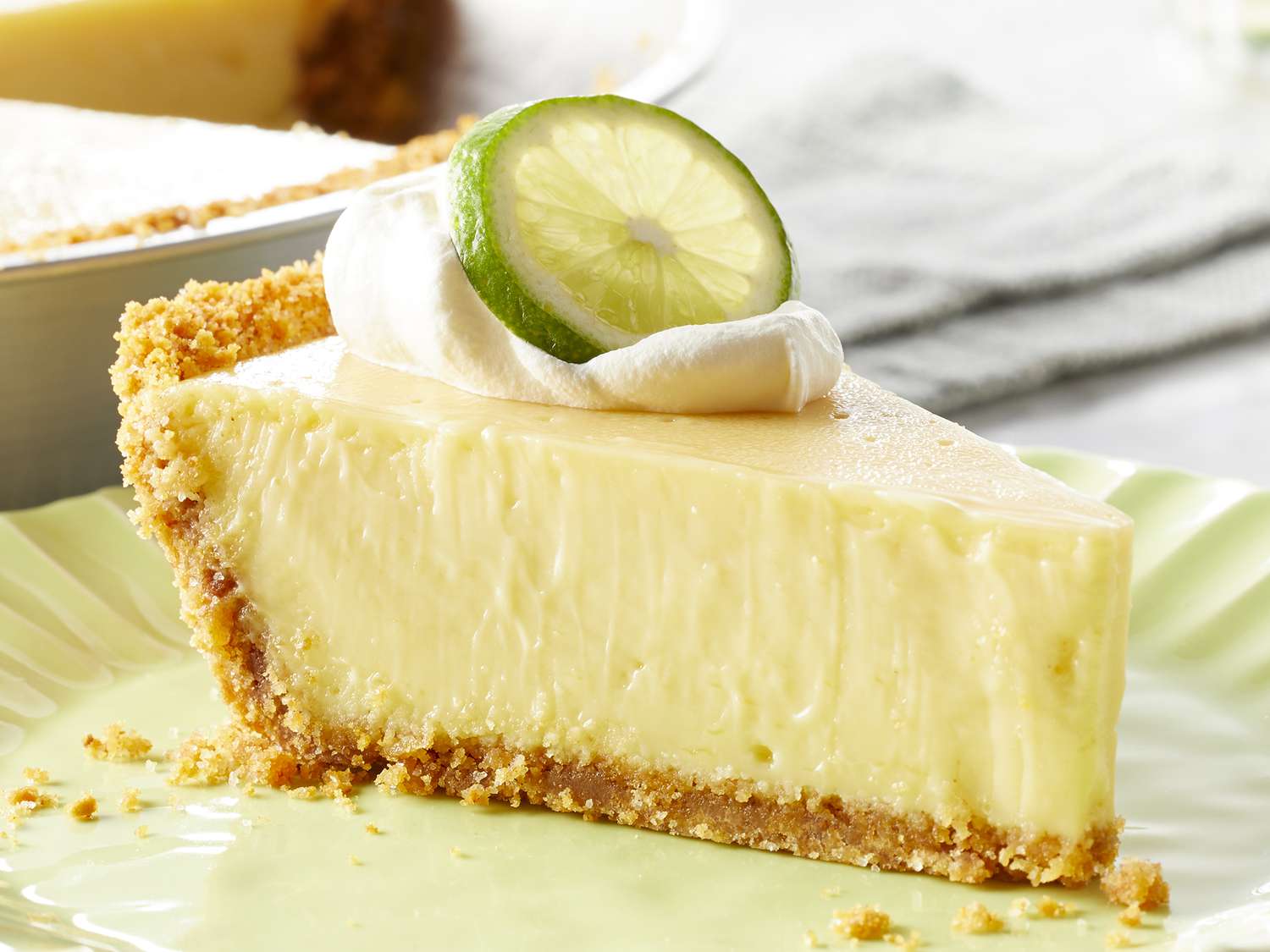A chilled slice of key lime pie with a pale yellow filling, graham cracker crust, and a small swirl of whipped cream on top, photographed on a bright kitchen table