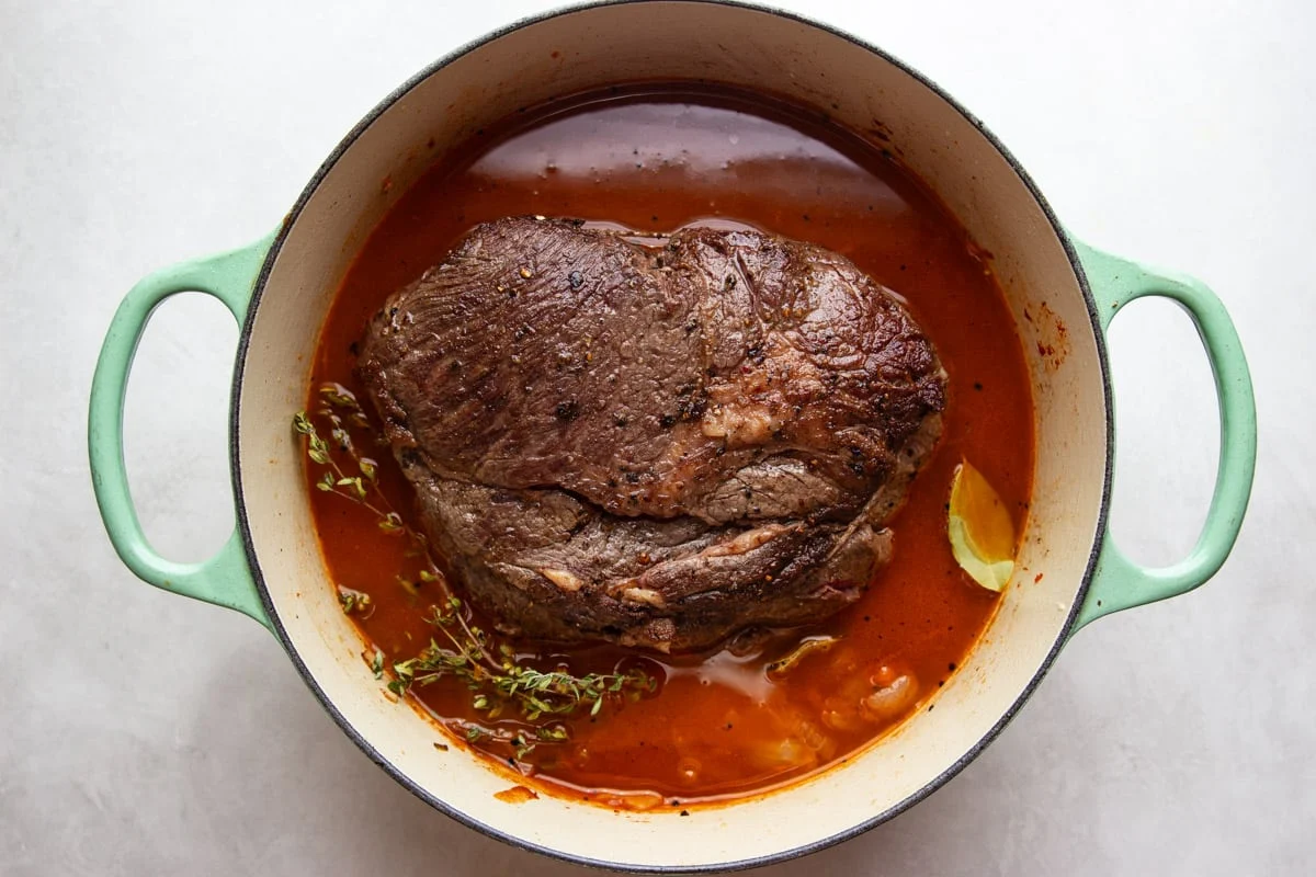 A chuck roast on a cutting board being seasoned with kosher salt and black pepper, hands visible, warm kitchen lighting