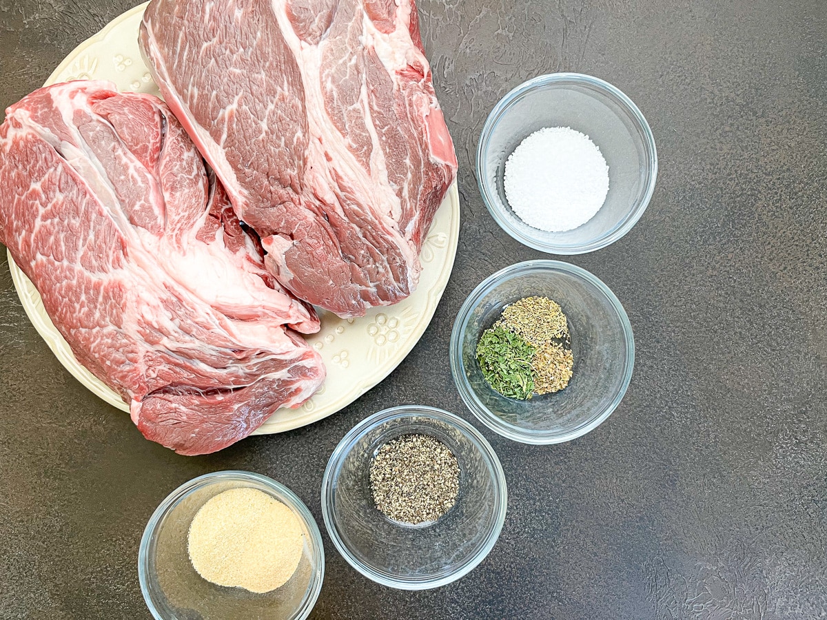 A chuck roast on a cutting board being seasoned with salt, pepper, and dried herbs, with small bowls of spices nearby