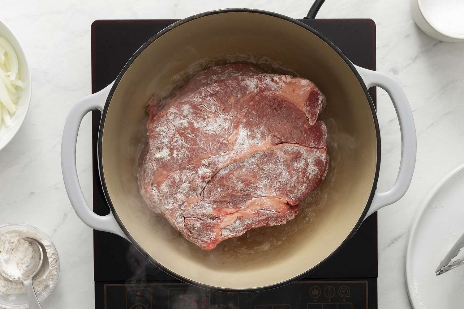 A chuck roast seasoned with salt and pepper being lightly coated with flour on a cutting board next to a skillet