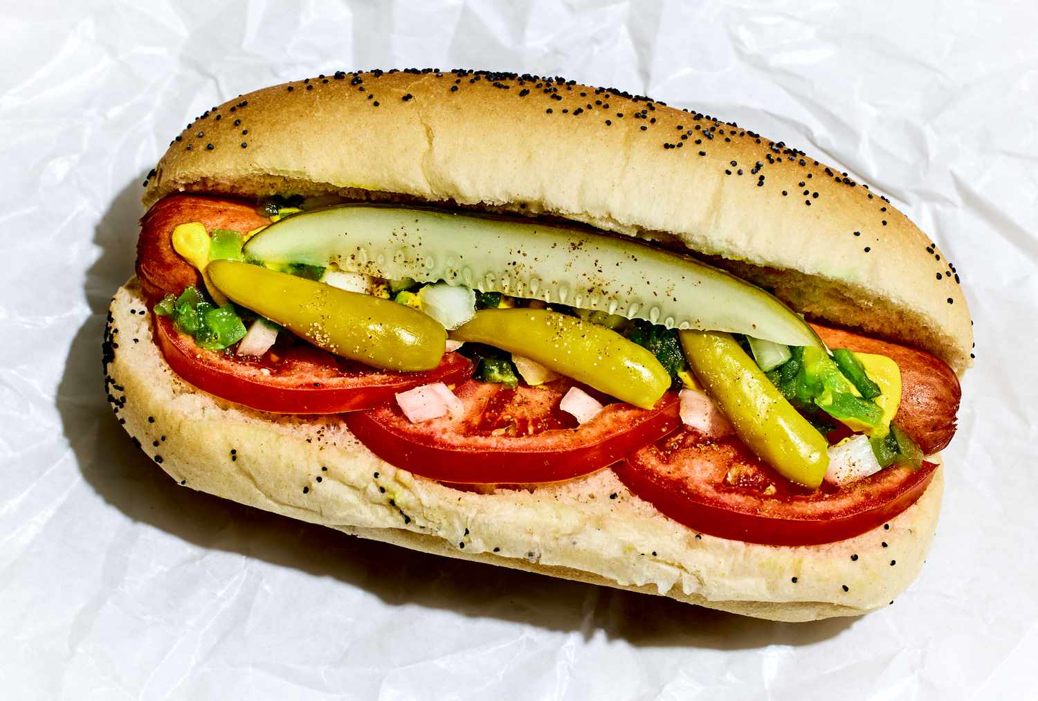 A classic Chicago-style hot dog in a poppy seed bun topped with yellow mustard, neon-green relish, chopped white onion, tomato wedges, a pickle spear, sport peppers, and a sprinkle of celery salt on a simple kitchen counter