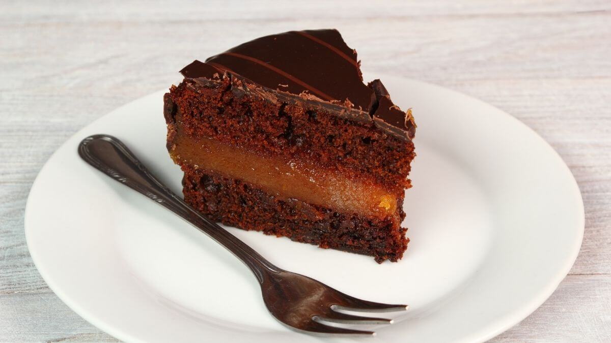 A classic Sachertorte with a mirror-like dark chocolate glaze on a cake stand, one slice cut to show the apricot jam layer, on a warm-lit kitchen table