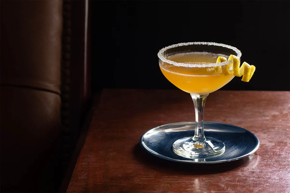 A classic Sidecar cocktail in a chilled coupe glass with a half sugar rim and a curled lemon twist on a neutral stone countertop under warm kitchen lighting