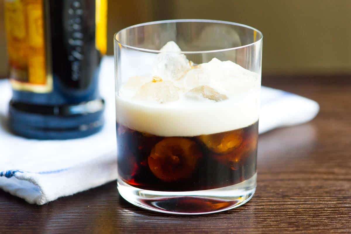 A classic White Russian cocktail in a clear rocks glass with ice, creamy swirls mixing into coffee liqueur and vodka, set on a warm kitchen counter with soft evening light