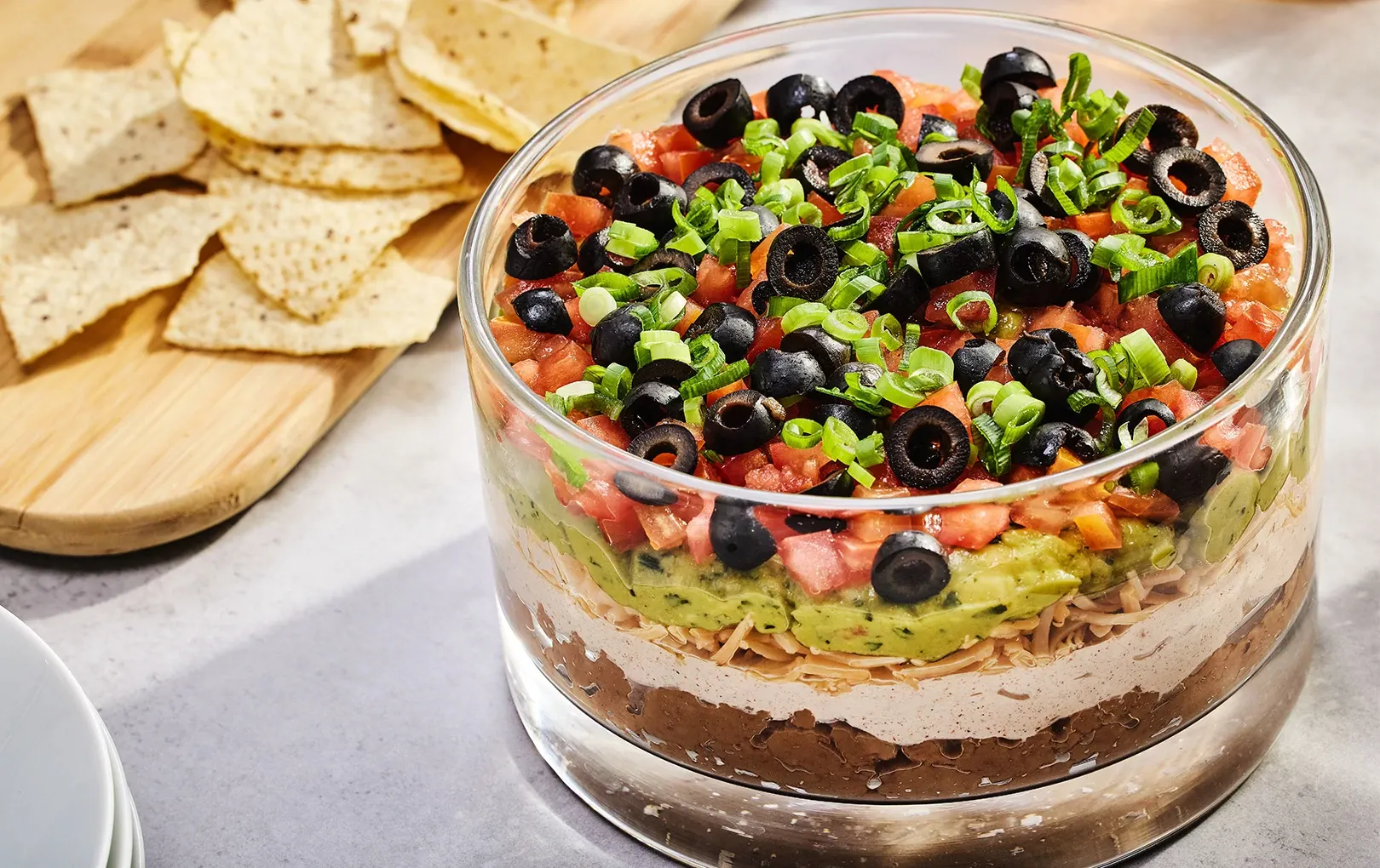 A clear glass baking dish filled with seven layer dip showing distinct layers of refried beans, guacamole, sour cream, salsa, shredded cheese, and fresh toppings on a bright kitchen counter