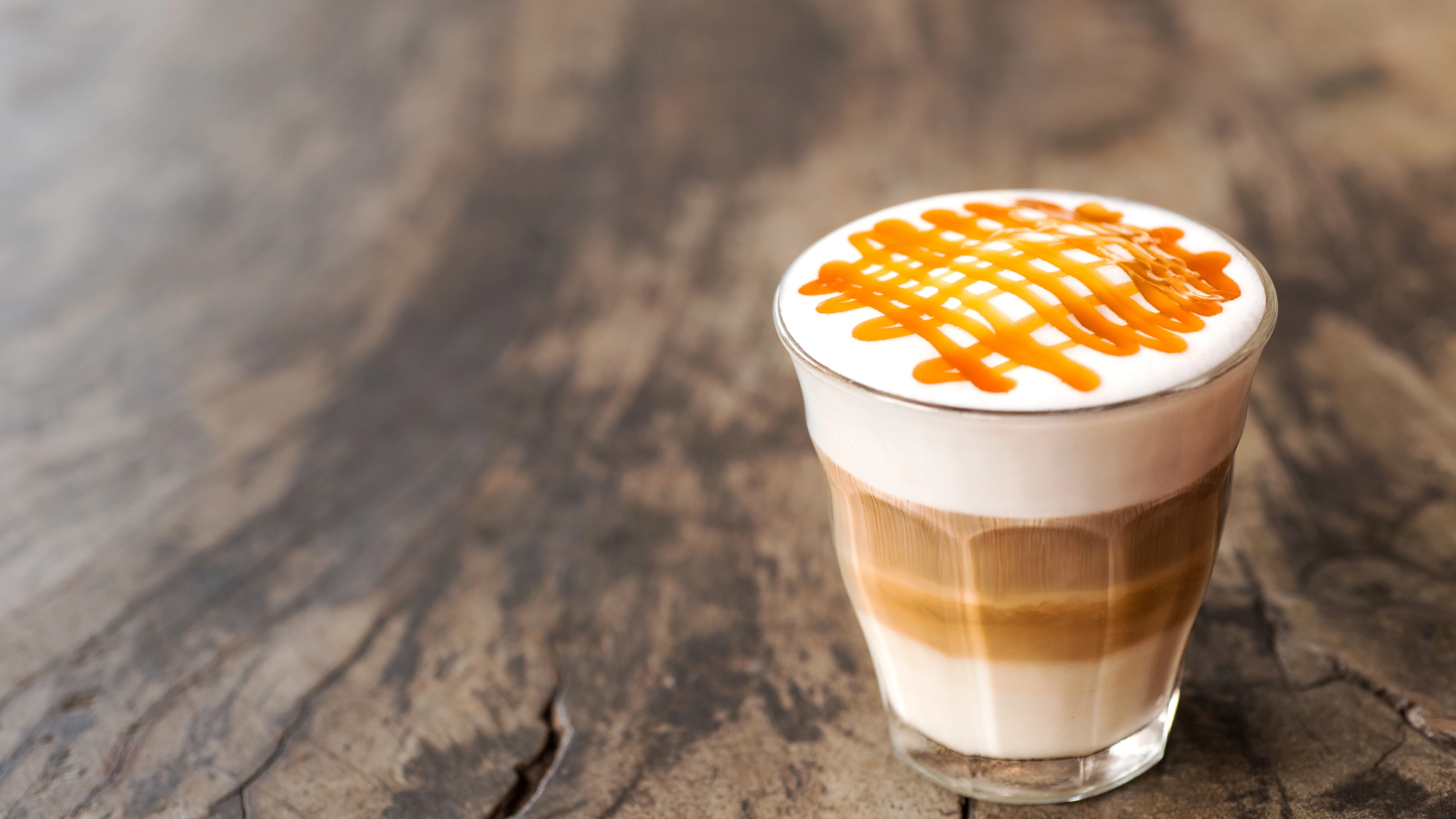 A clear glass caramel macchiato with layered espresso on top of foamed milk and caramel drizzle, sitting on a light stone kitchen counter in soft morning light