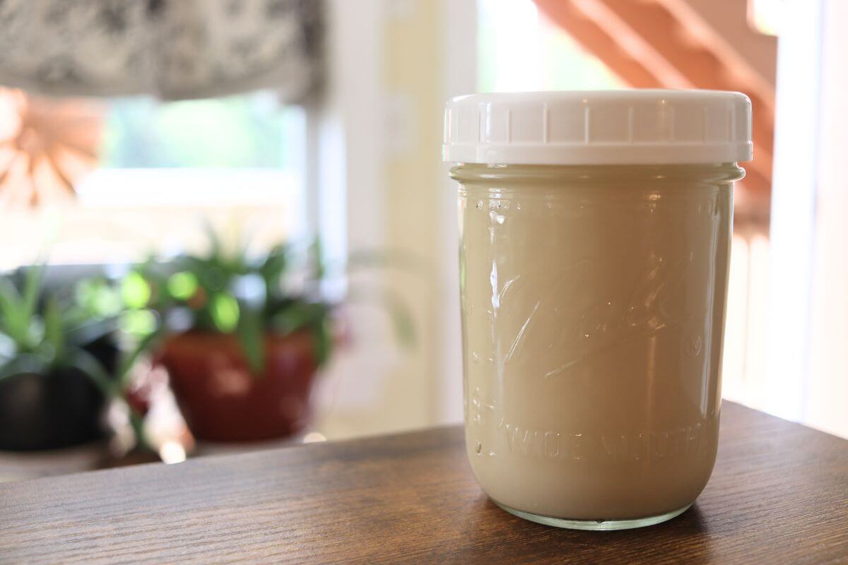A clear glass jar filled with creamy homemade oat milk on a bright kitchen counter with a small bowl of rolled oats nearby