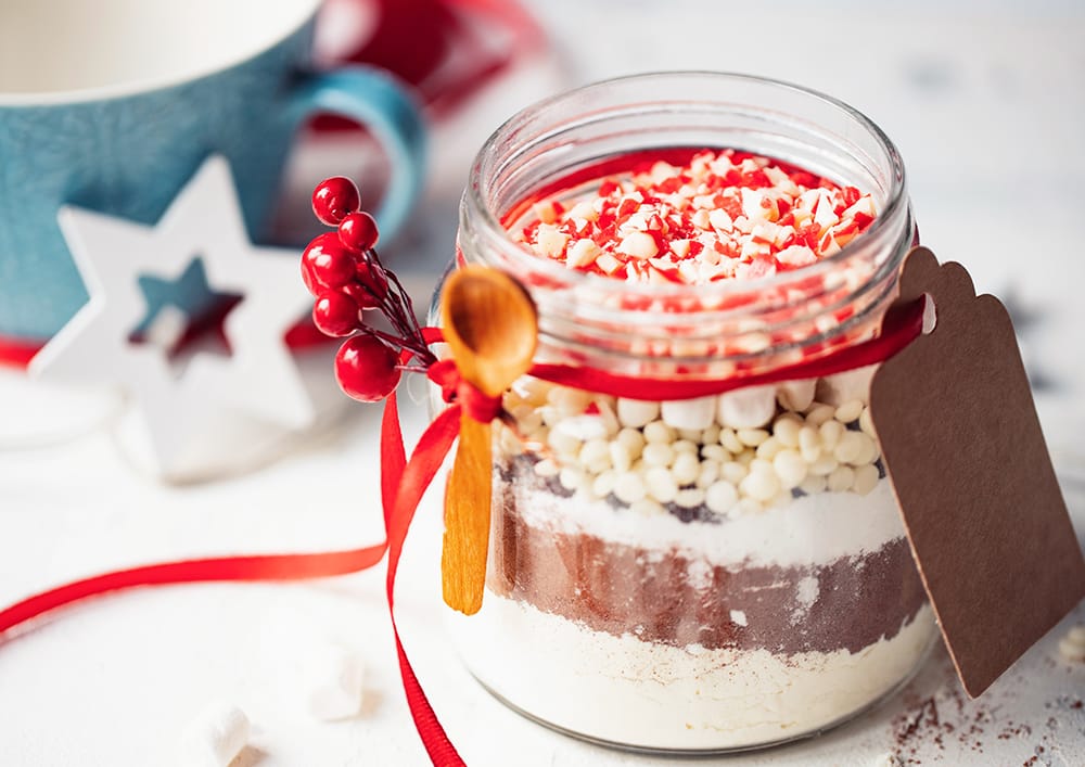 A clear glass jar filled with homemade hot cocoa mix on a warm kitchen counter, with a spoon and a small bowl of cocoa powder nearby