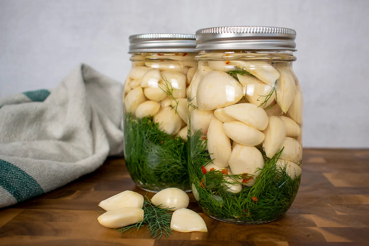 A clear glass jar filled with pickled garlic cloves in a pale vinegar brine with black peppercorns and a bay leaf on a bright kitchen counter
