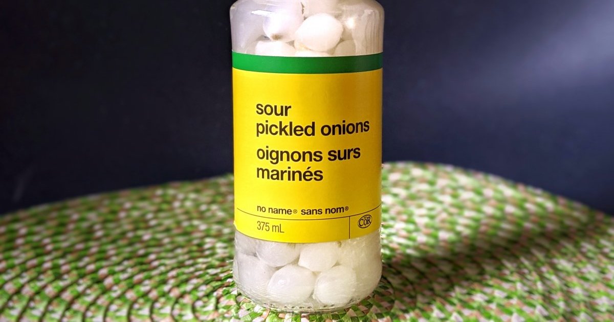 A clear glass jar filled with pickled pearl onions in golden brine with garlic cloves and peppercorns on a warm kitchen counter